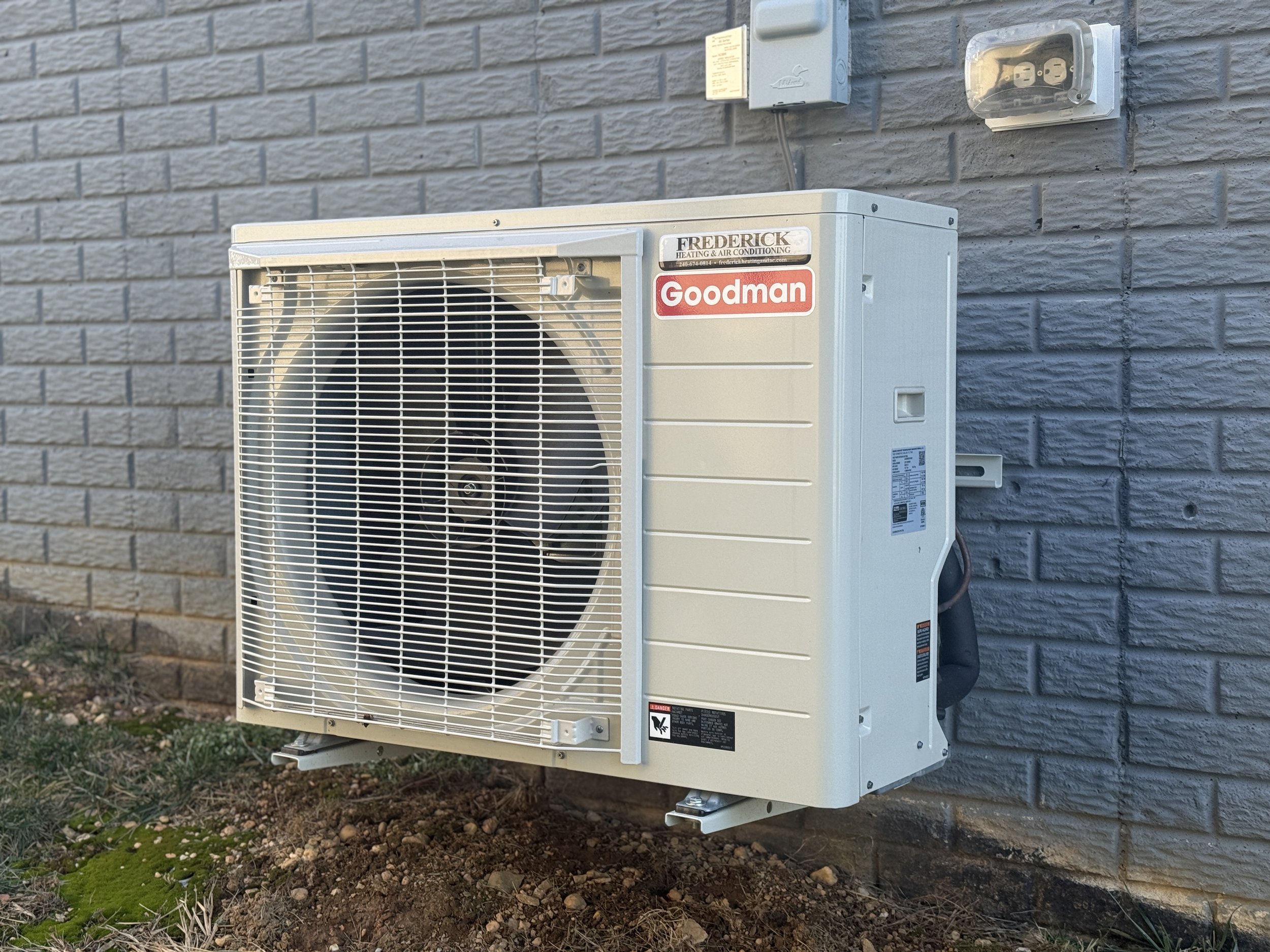 Outdoor HVAC unit attached to a gray brick wall next to a small patch of soil and plants.