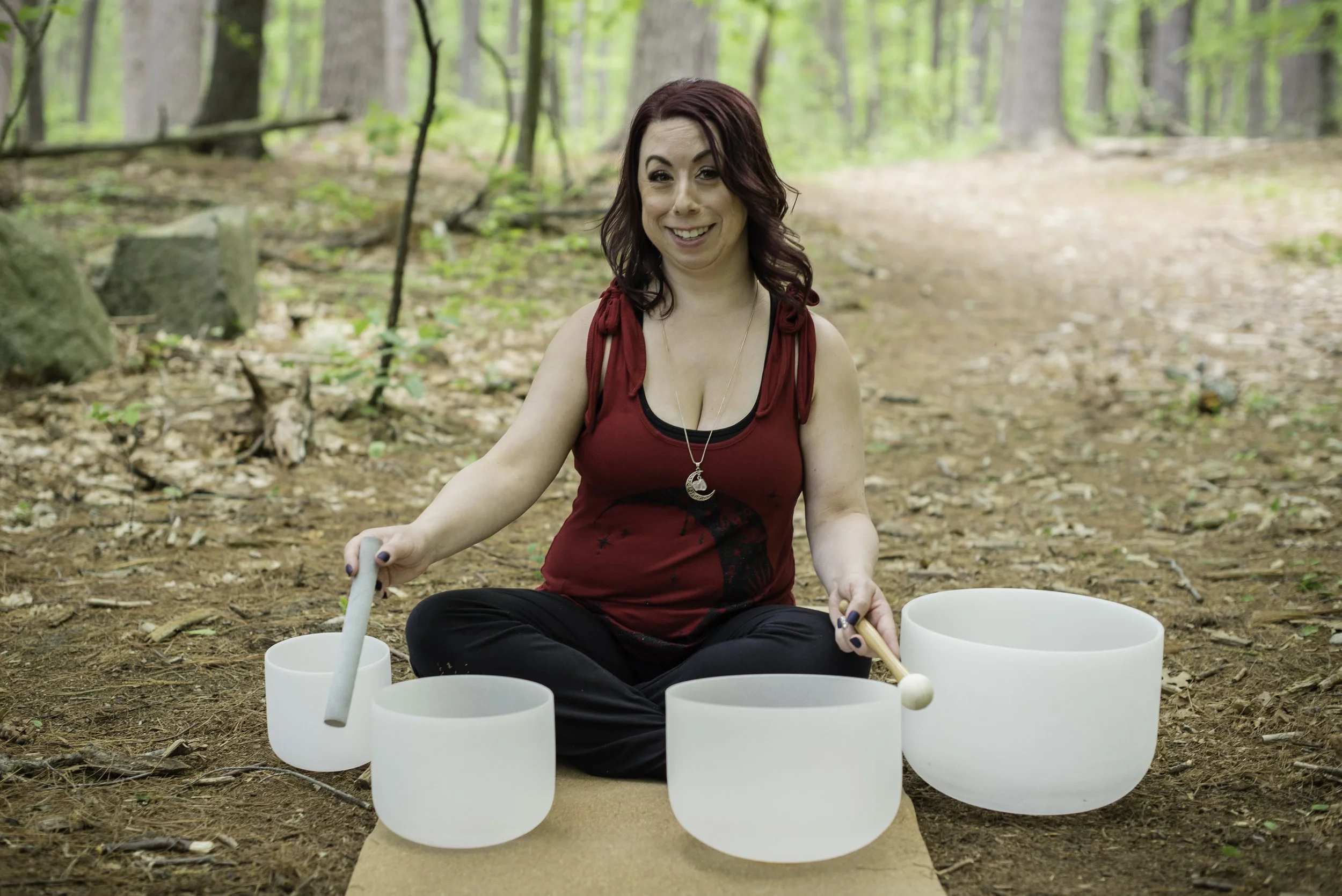 A woman with shoulder-length red hair smiling while sitting cross-legged on the ground in a forest, playing outdoors with white crystal singing bowls.