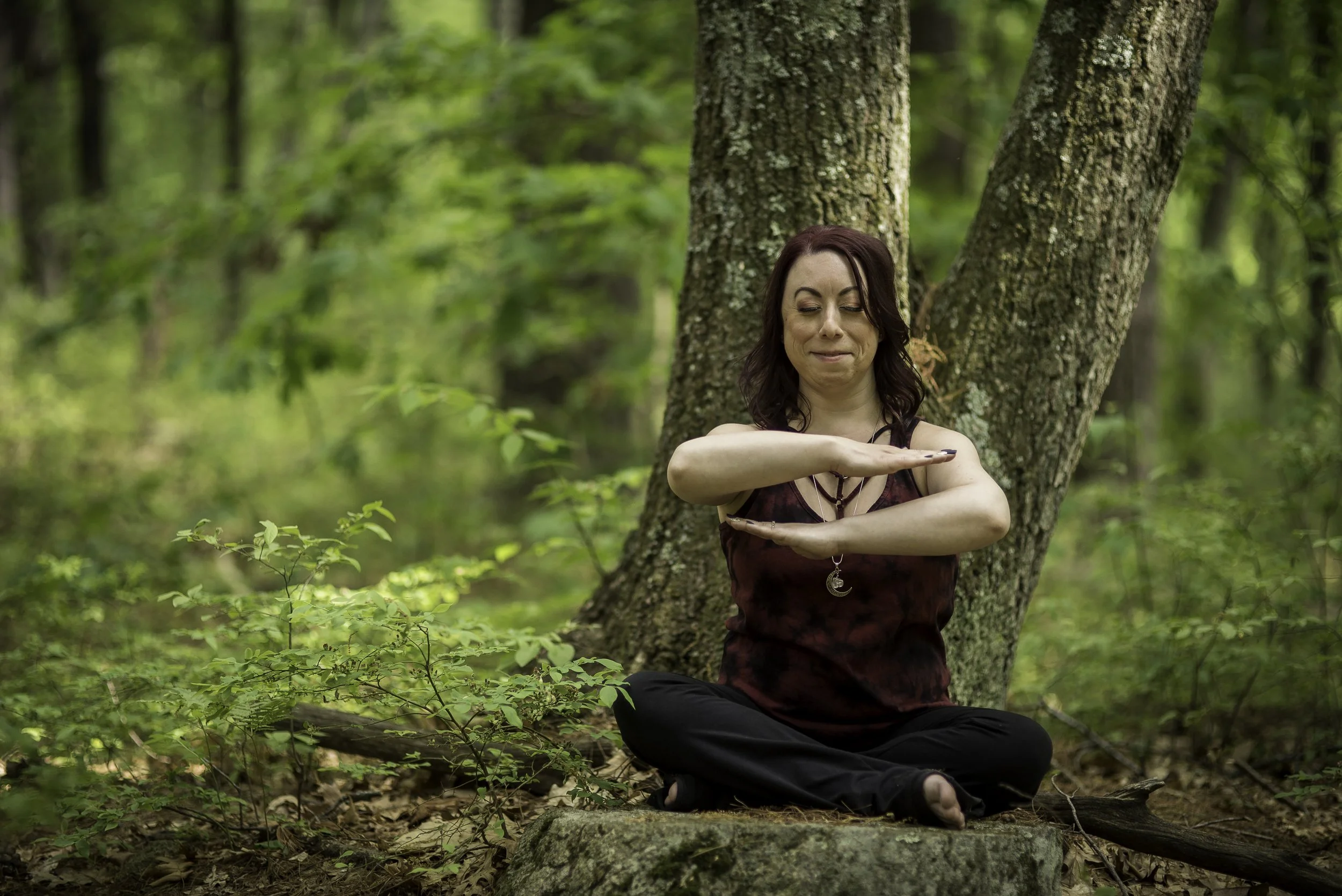 A woman with blonde long hair tied up meditation with beige pants red socks and black tank top with left hand on heart and right hand on stomach with green plant behind her