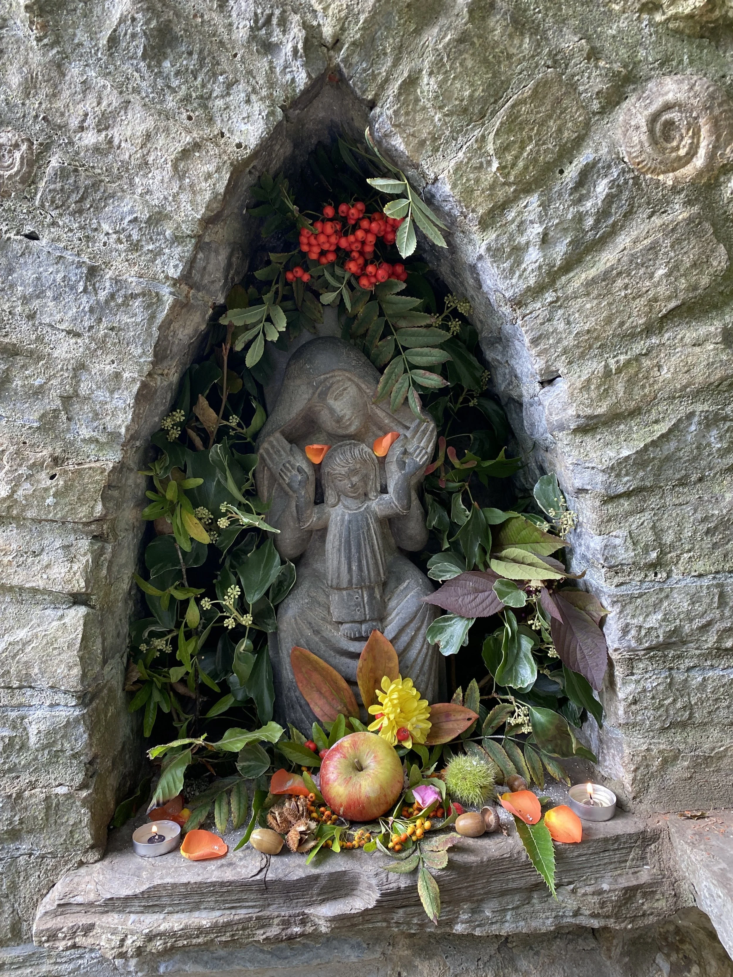 A religious shrine with a stone statue of Mary Magdalene surrounded by green foliage, red berries, an apple, candles, and floral decorations in a stone nook.  Glastonbury retreat Chalice Well