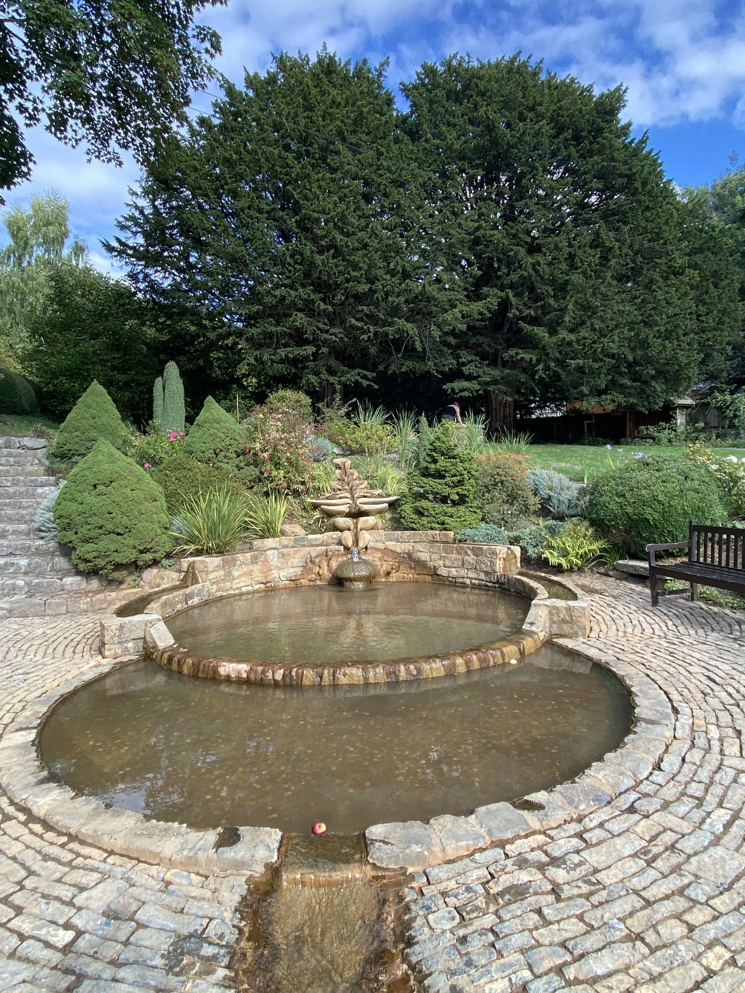 A landscaped garden with a stone fountain in the center, two tiered with water cascading down, surrounded by lush green bushes, small trees, and colorful plants. 
Glastonbury retreat. Chalice Well