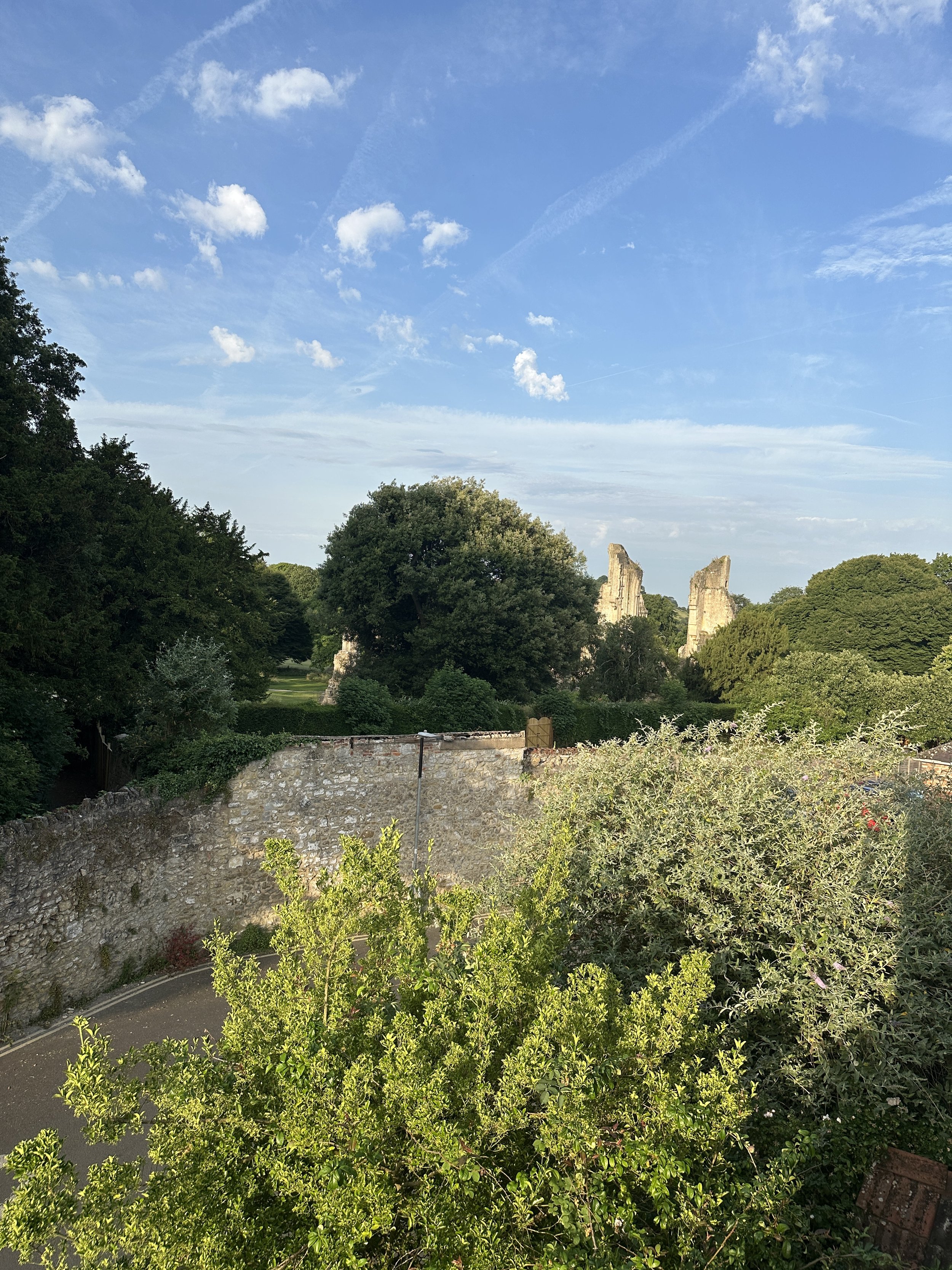 Glastonbury retreat. A scenic view of a garden with trees, shrubs, and a stone wall, with ruins of tall stone structures in the background under a partly cloudy blue sky.
