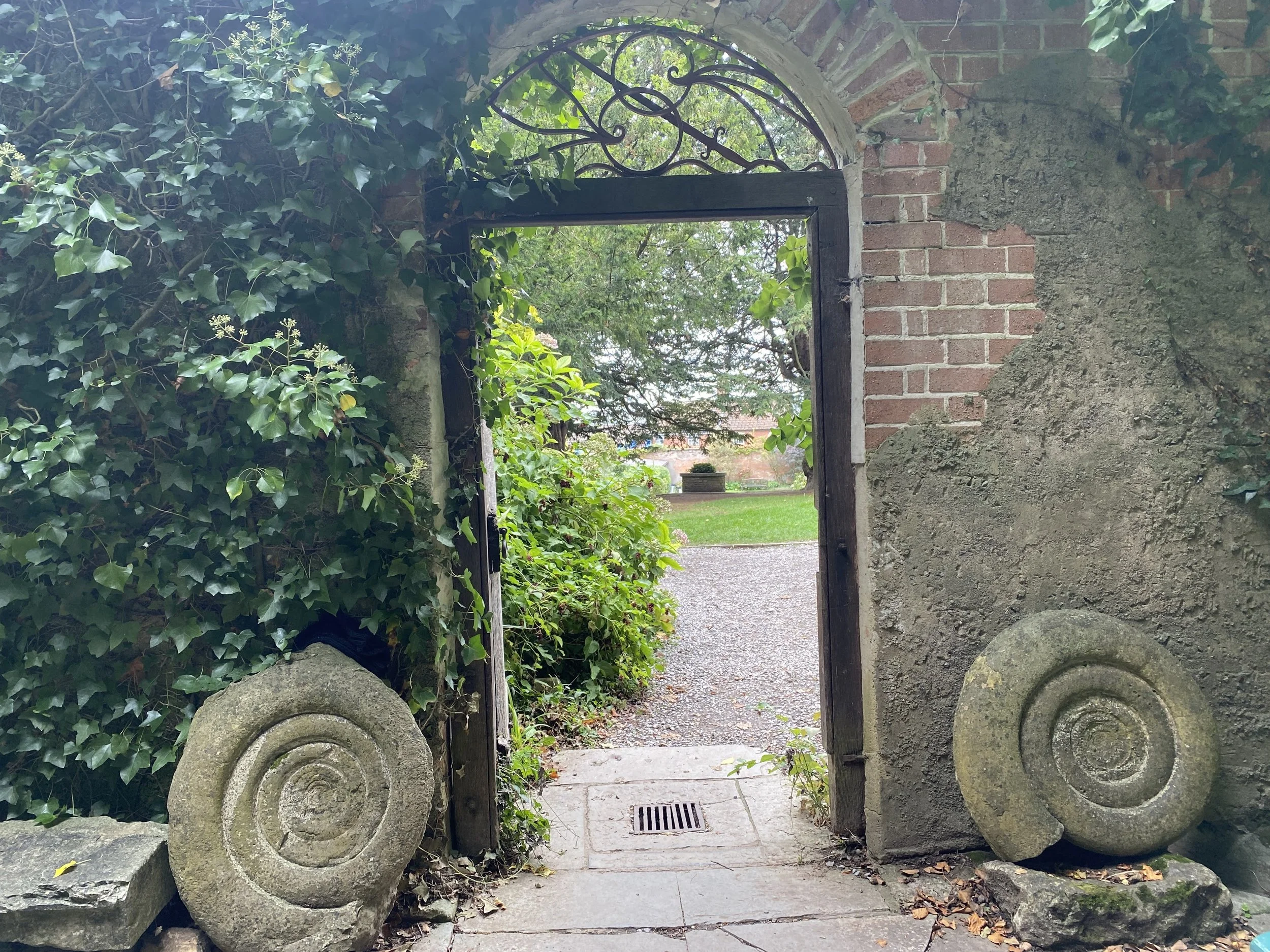 
Glastonbury retreat. Chalice Well An arched garden gate made of wood and wrought iron, surrounded by ivy and stone sculptures, leading to a pathway and a grassy area with trees in the background.