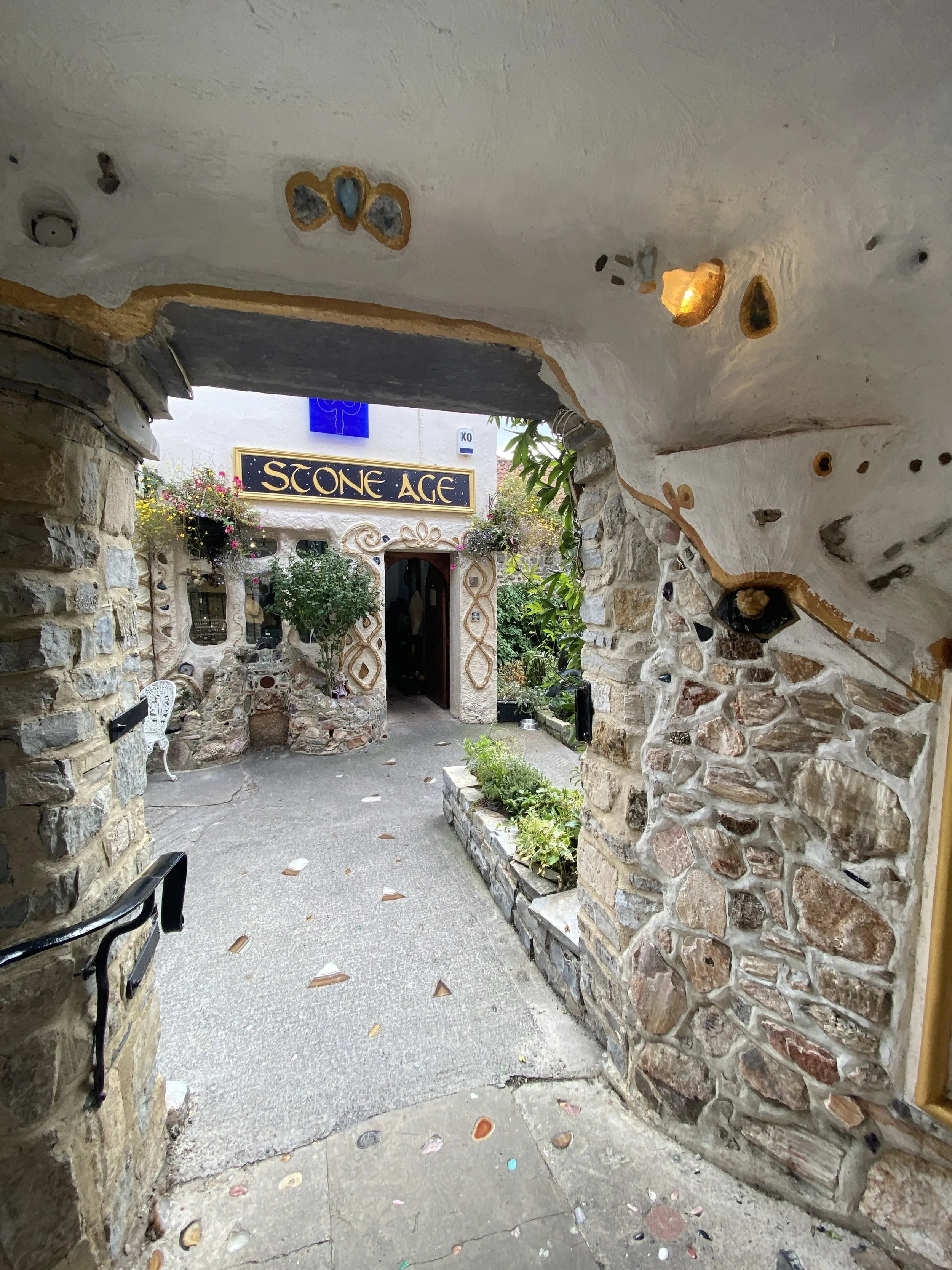 	
Glastonbury retreat High street. s Entrance to a building called 'Stone Age' with a stone and stucco exterior, surrounded by greenery and flowers.