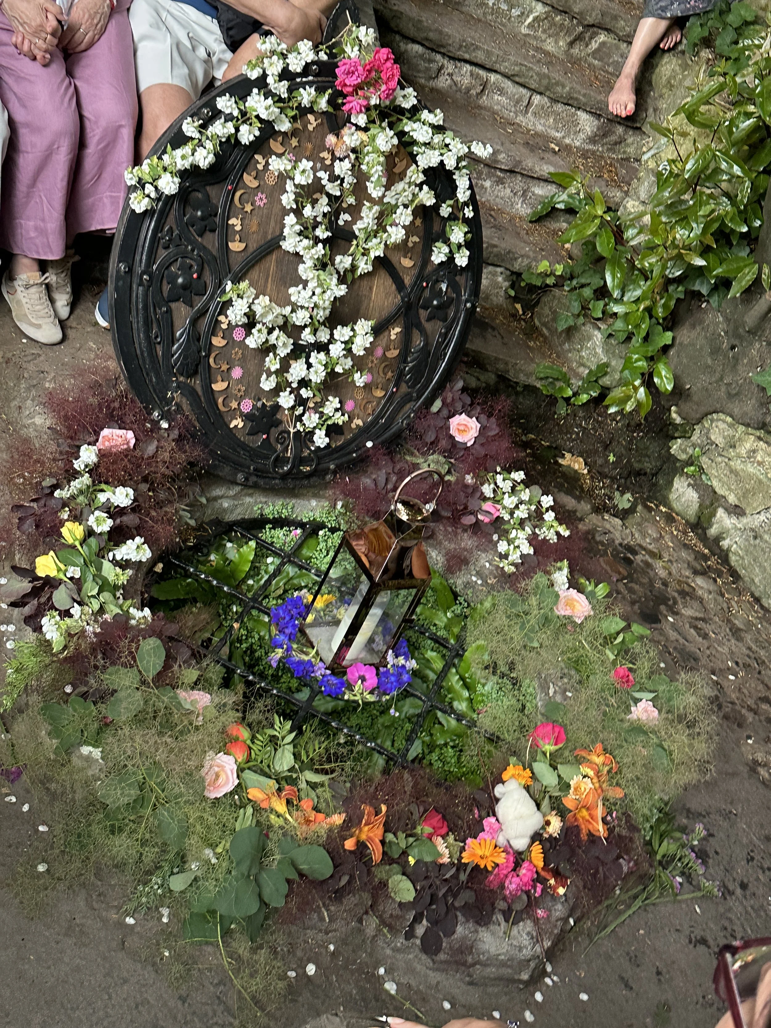 Glastonbury retreat Chalice Well. A memorial with flowers, candles, and a small stuffed animal, placed at a stone wall with people gathered around.