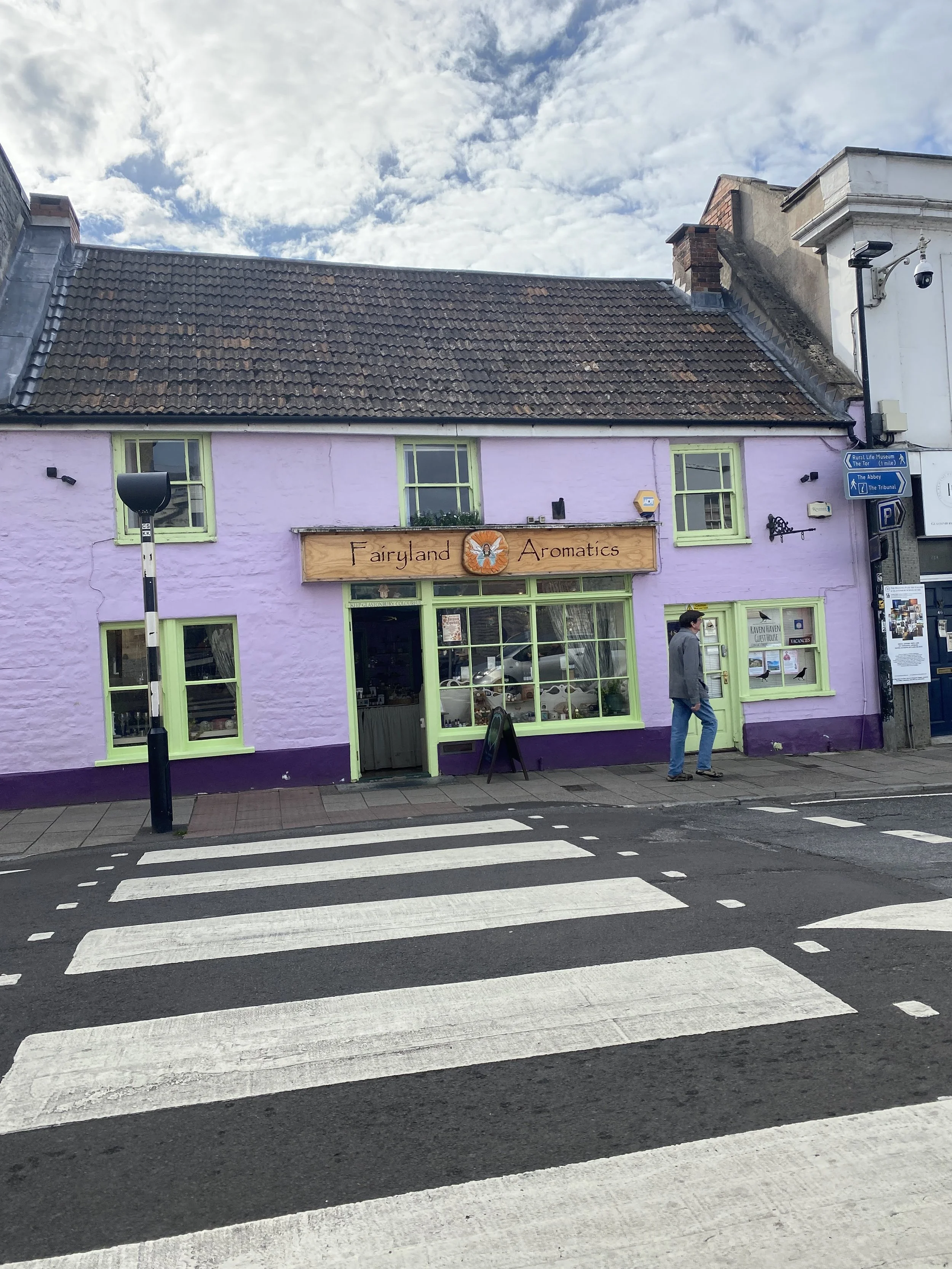 A purple building with green window frames and a sign that reads 'Fairyland Aromatics'. A person is walking on the sidewalk in front of the building, which is located across a crosswalk on the street. Glastonbury retreat High street. shopping