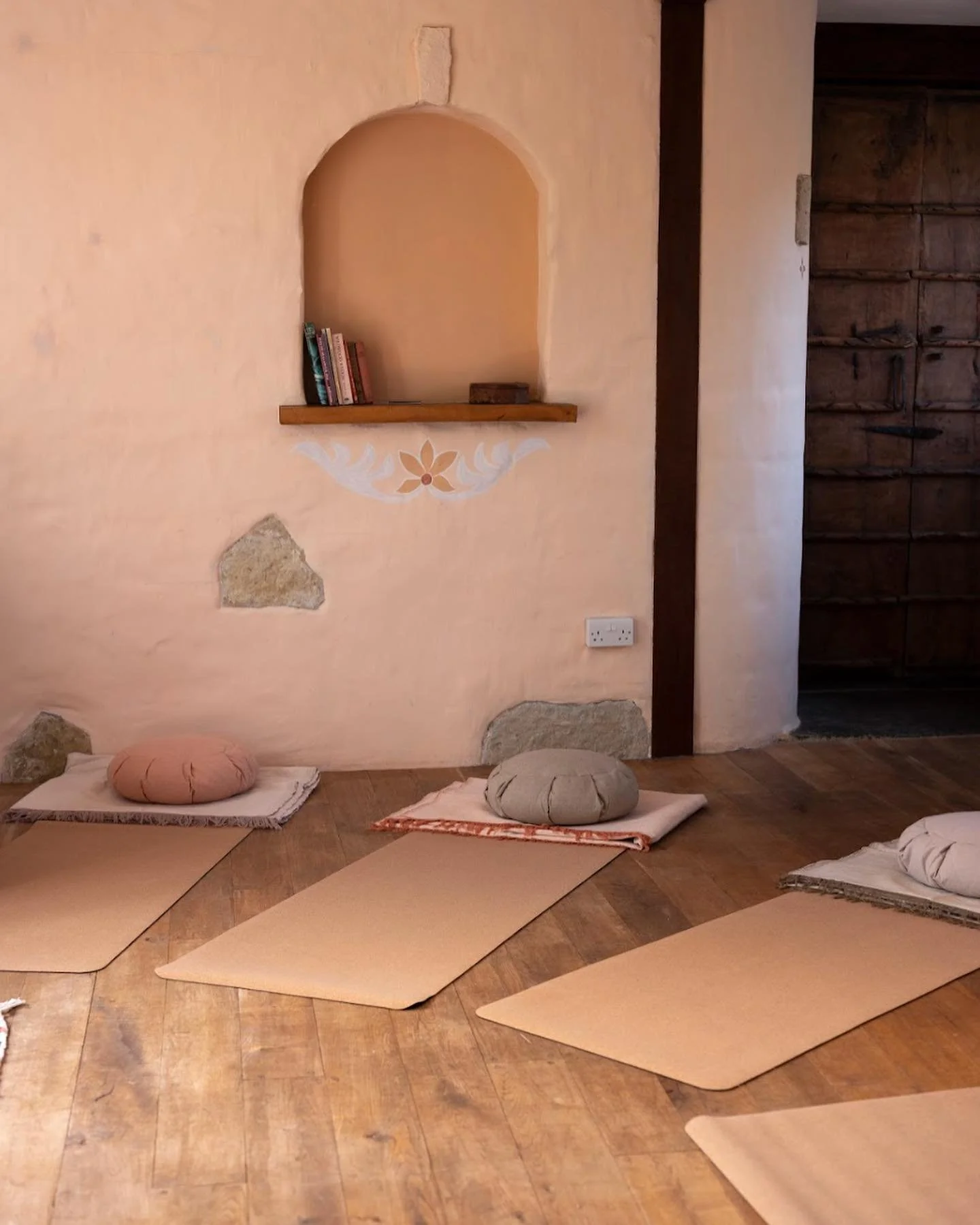 Glastonbury retreat Apothecary Gardens Three yoga mats with cushions on top, placed on a wooden floor in a room with a peach-colored wall that has decorative stones and a small shelf with books.