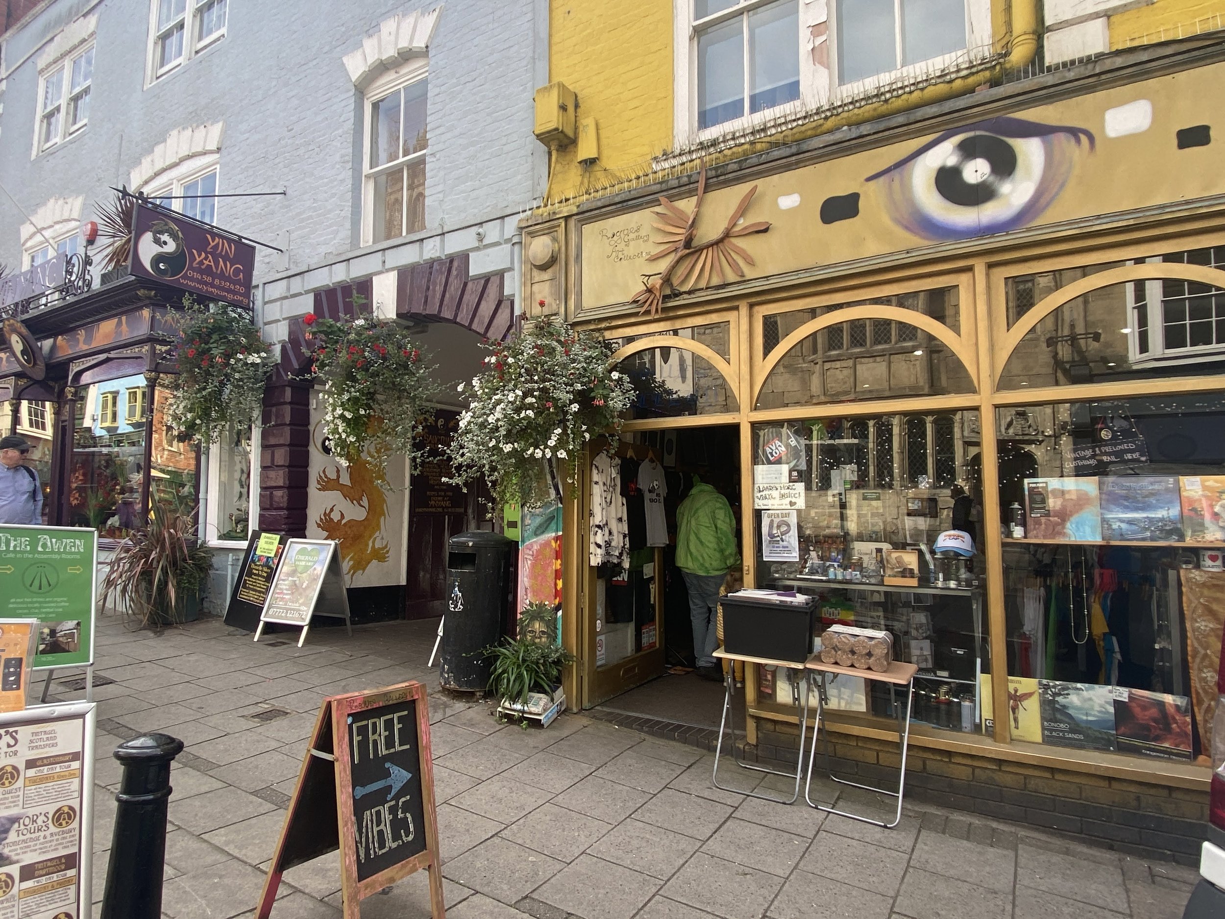 Glastonbury retreat High street. shopping Street view of a storefront with graffiti art, including a large eye painted above the window, and signs advertising free vibes. There are hanging flower baskets and people walking nearby.