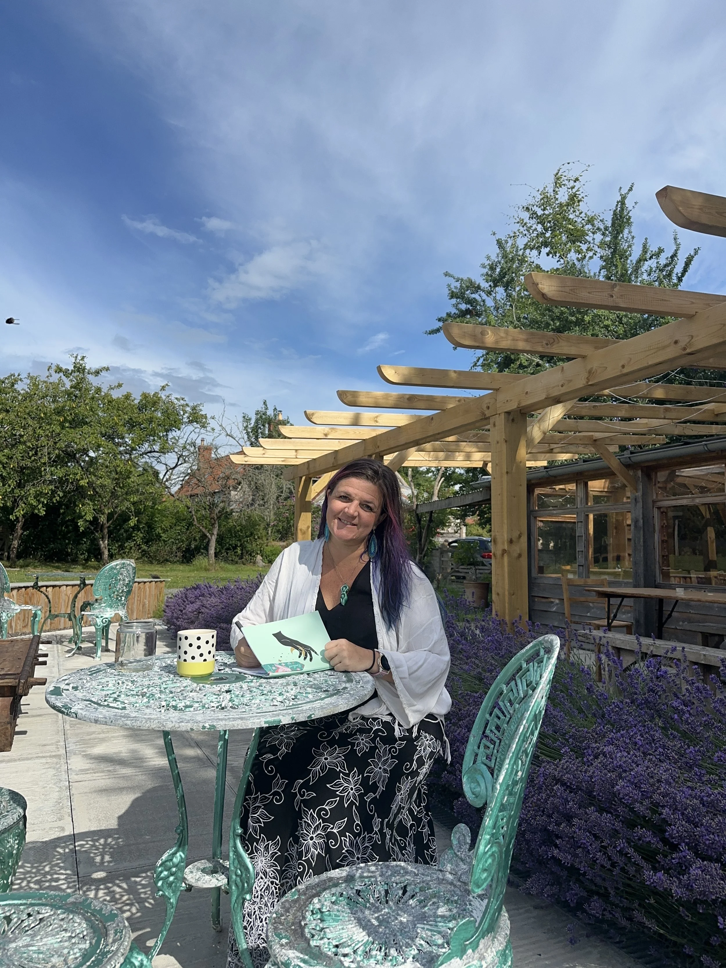 A woman sitting at a round table outdoors, holding a card with a fish illustration in her hand, surrounded by purple flowers.  Glastonbury retreat  Apothecary Garden