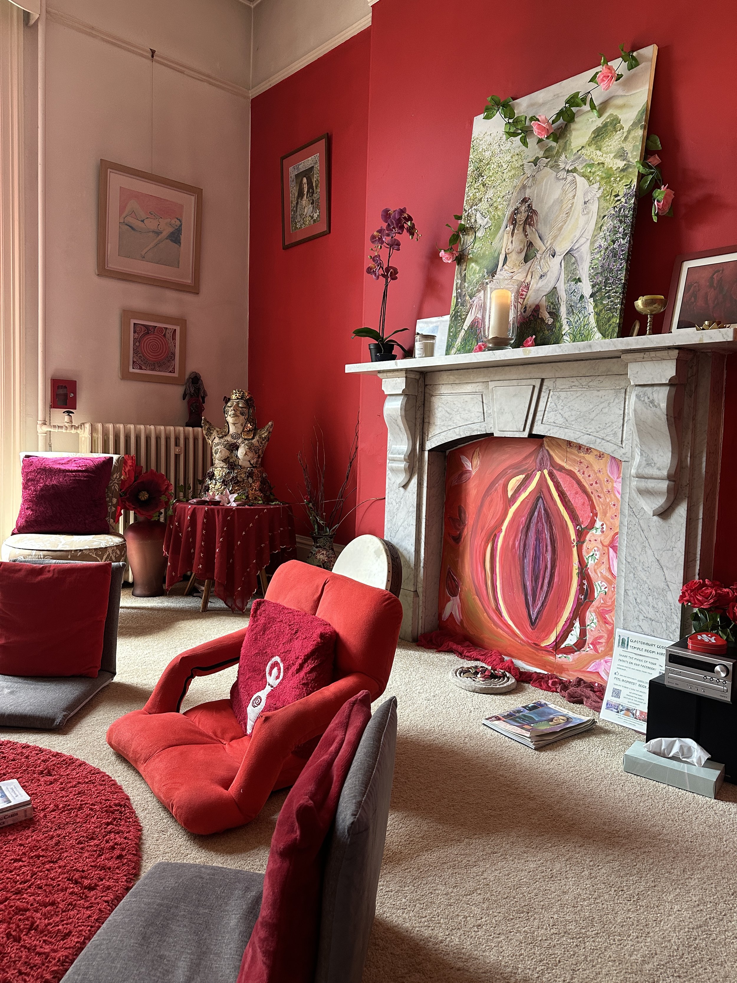 Glastonbury retreat Goddess House. Living room with red and pink decor, featuring a red armchair with a pink pillow, a fireplace with pink artwork, an ornate sculpture on a small table, and various framed pictures on the walls.