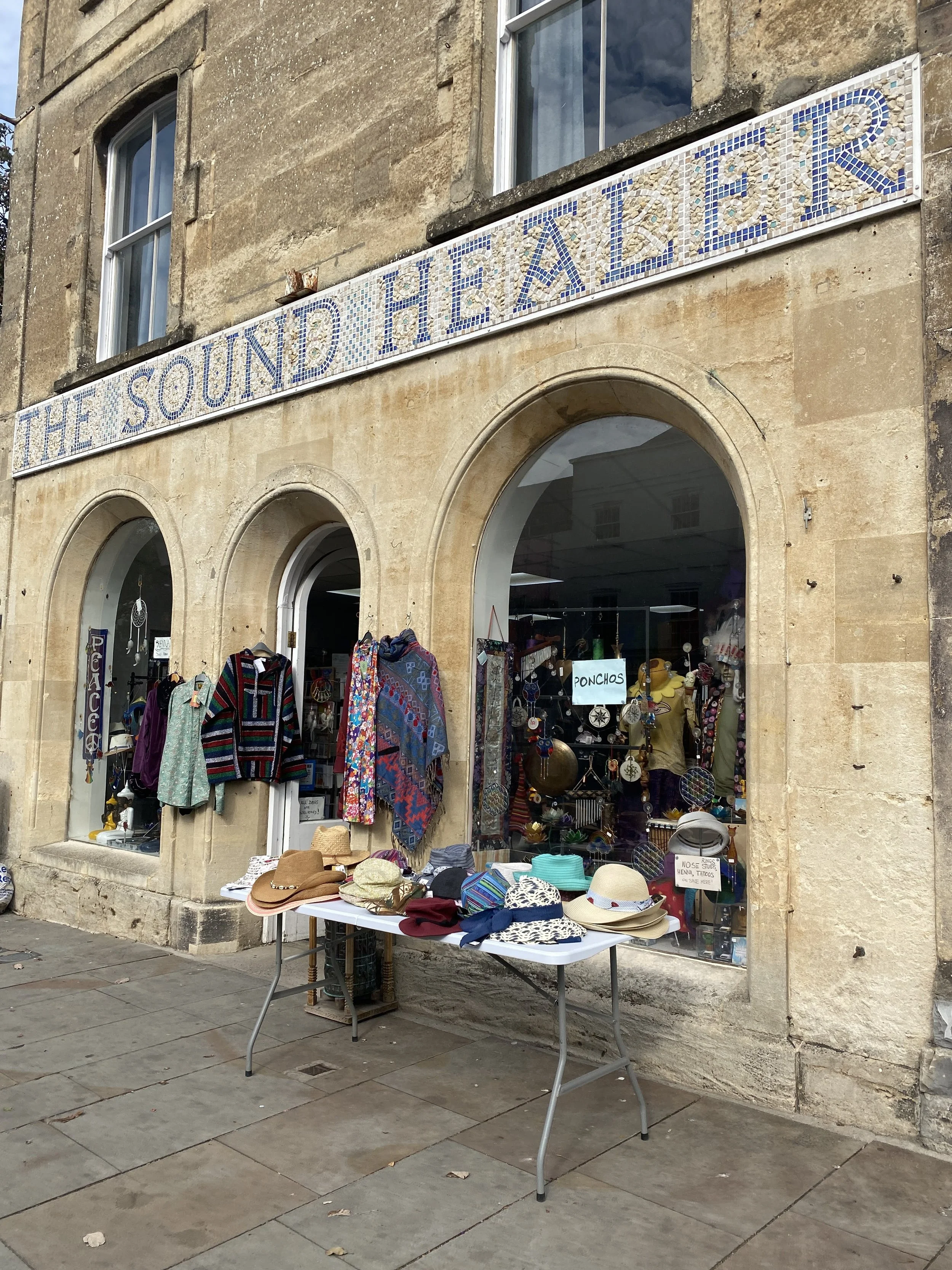 A store named 'The Sound Healer' with items for sale outside, including hats, scarves, ponchos, and jewelry, in front of a stone building with arched windows. Glastonbury retreat High street. shopping