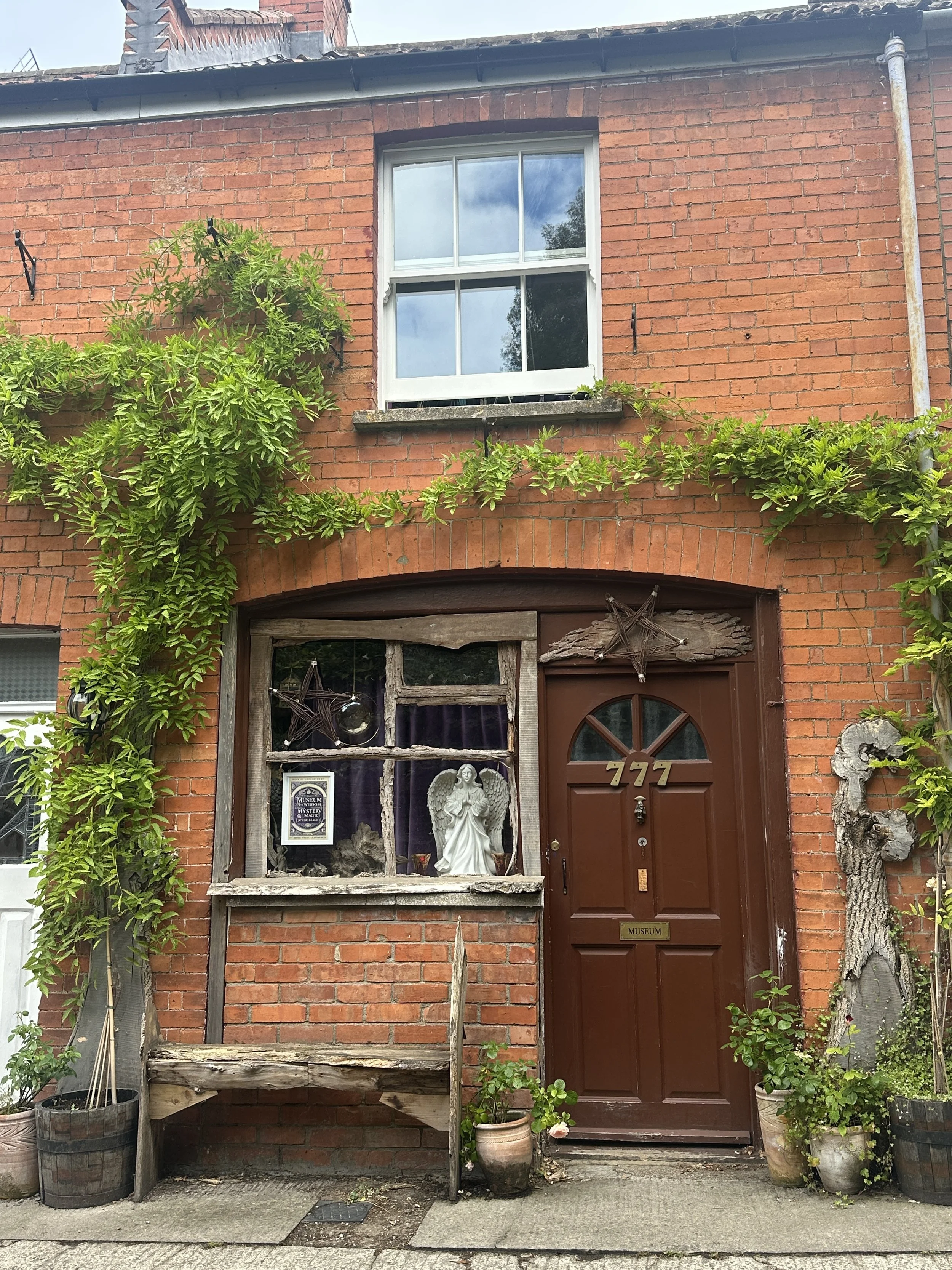 
Glastonbury retreat. Brick house with a window above a red door, decorated with star-shaped ornaments, a statue of an angel, and various potted plants and greenery around the entrance.