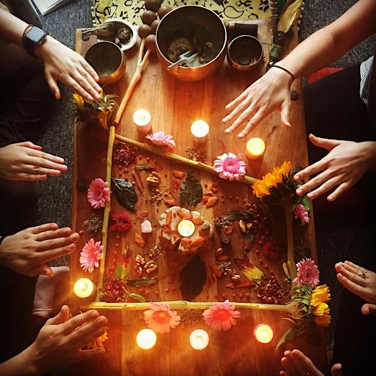 People gathered around a wooden table with flowers, candles, leaves, and small decorative objects, possibly for a spiritual or ritualistic activity.