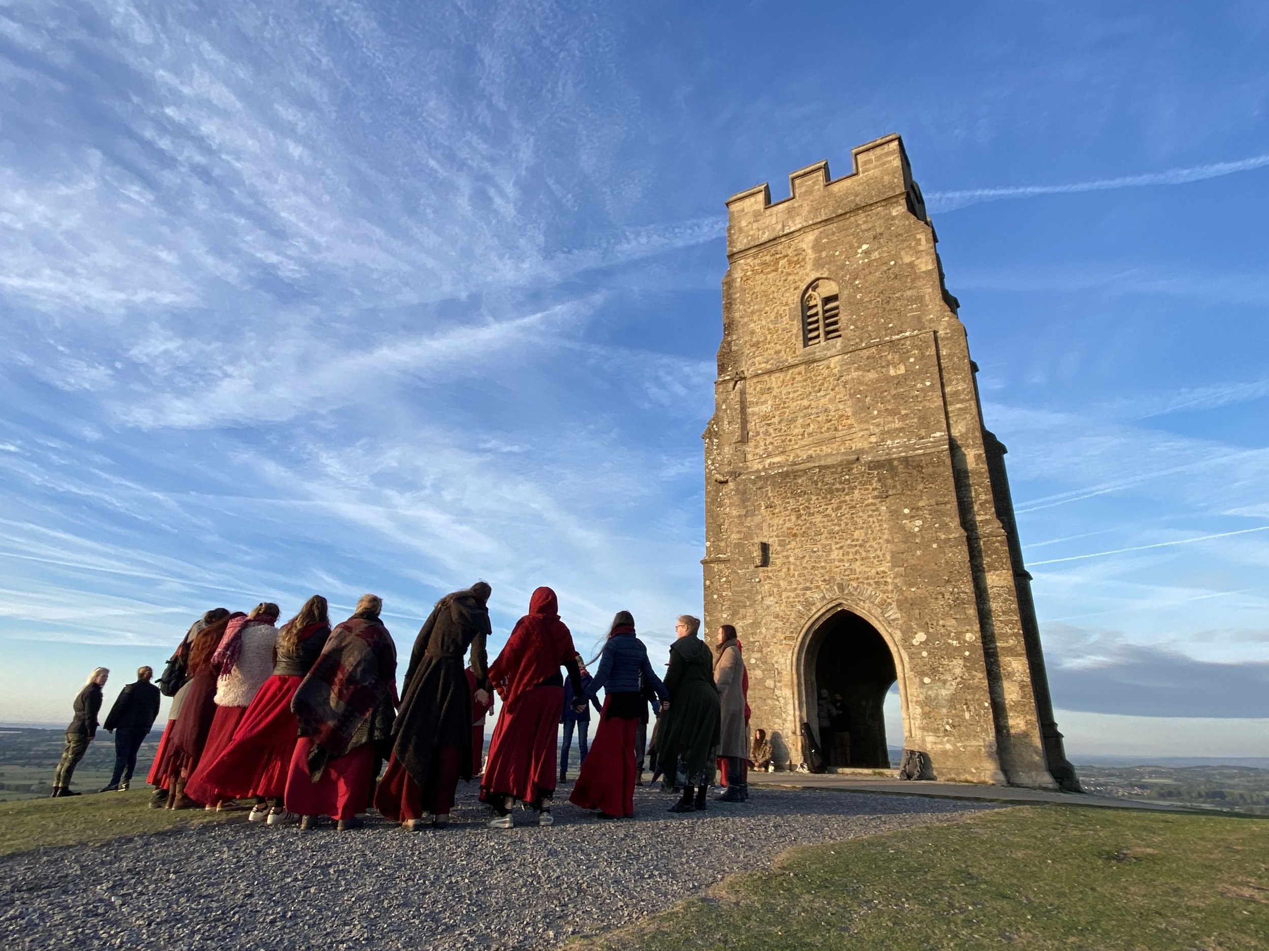 A group of people dressed in traditional Scottish clothing, including kilts and shawls, holding hands in a circle outside an ancient stone tower, with a scenic landscape and blue sky in the background.