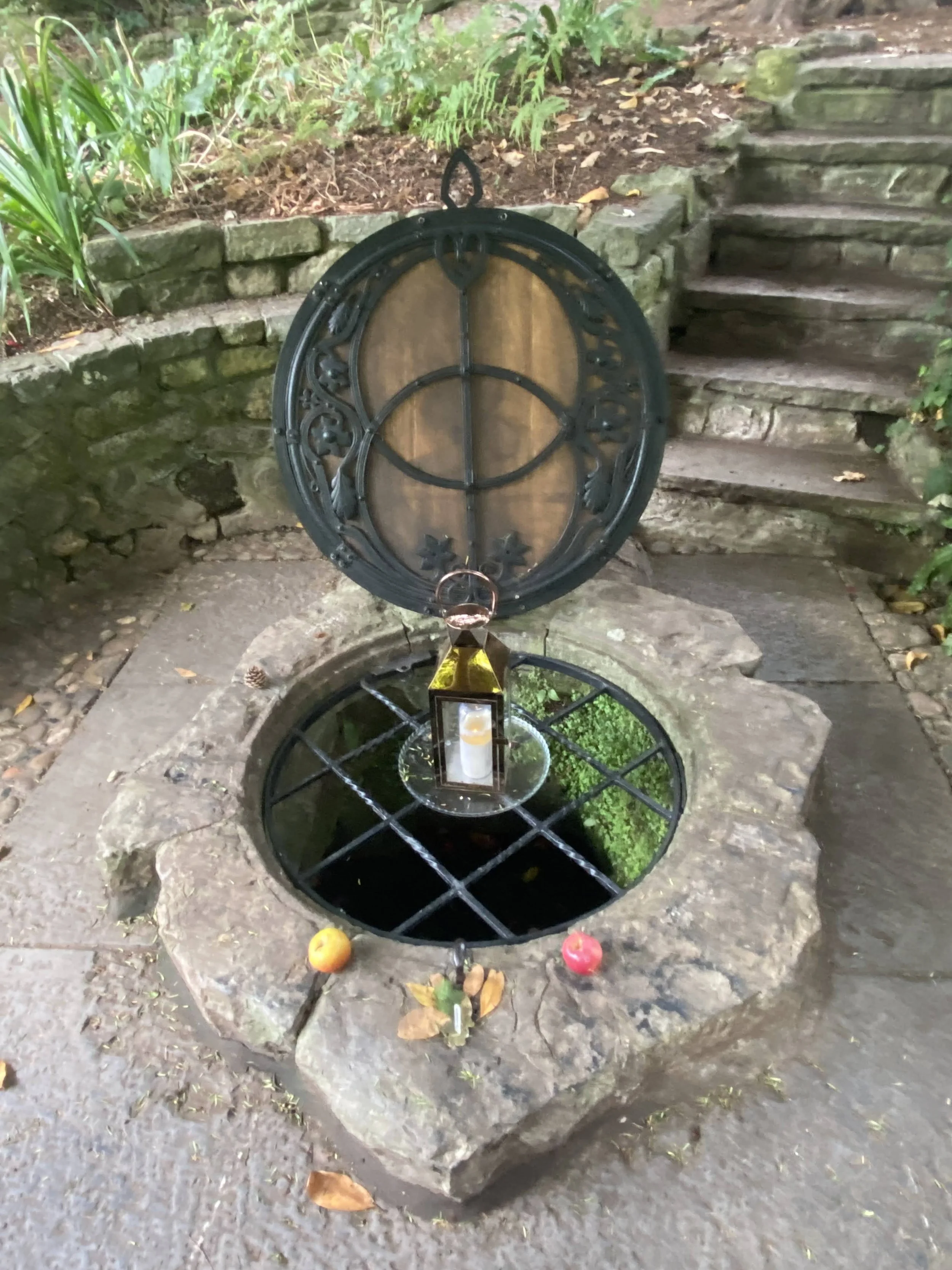 
Glastonbury retreat. Chalice Well A stone well with a metal cover and a lantern hanging above it, surrounded by a garden with plants and stone steps nearby.
