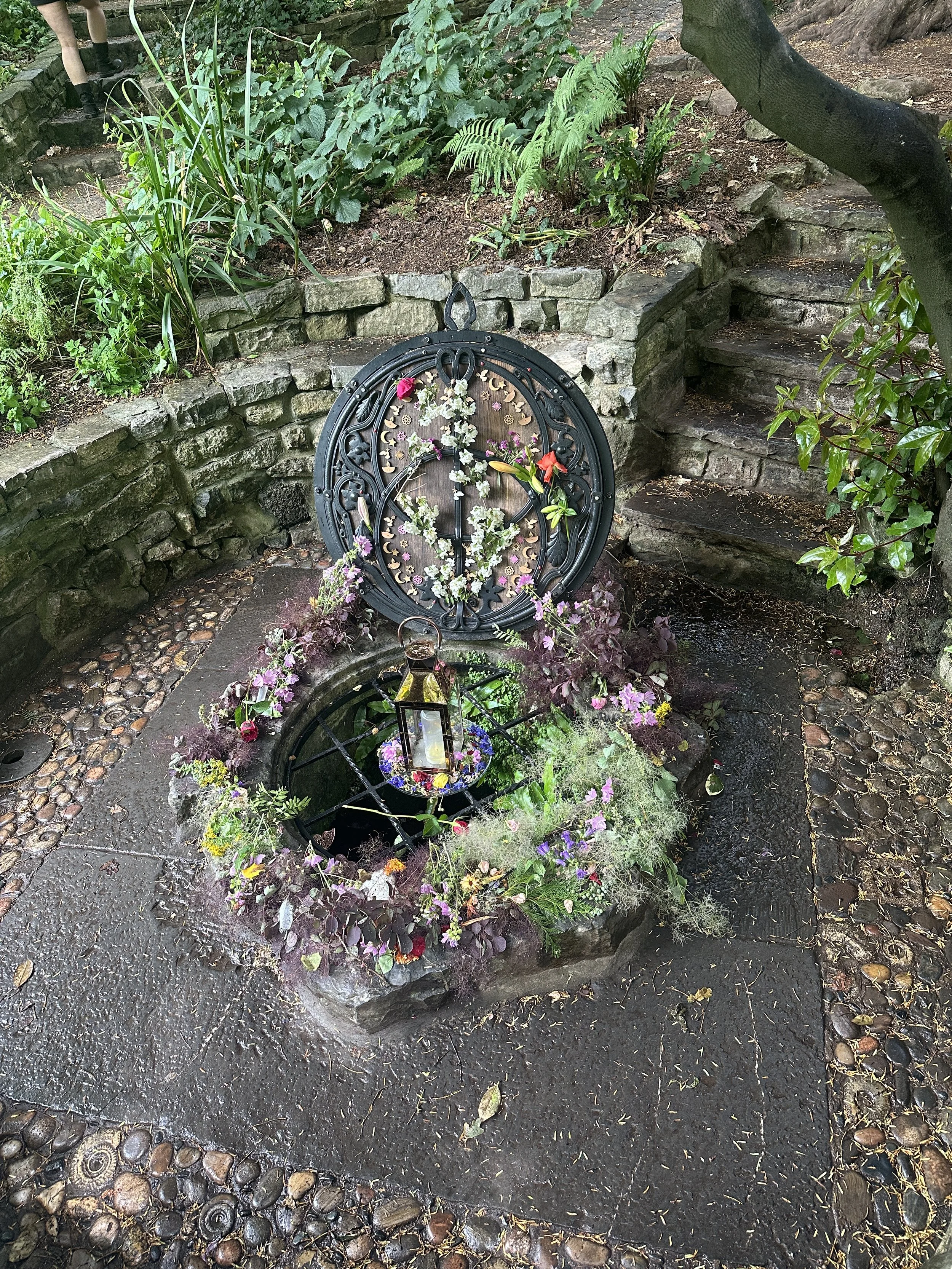 Glastonbury retreat Chalice Well. A decorative memorial with a circular metal frame adorned with flowers, placed over a small votive ground-level shrine surrounded by plants and greenery, with stone steps and garden walls in the background.