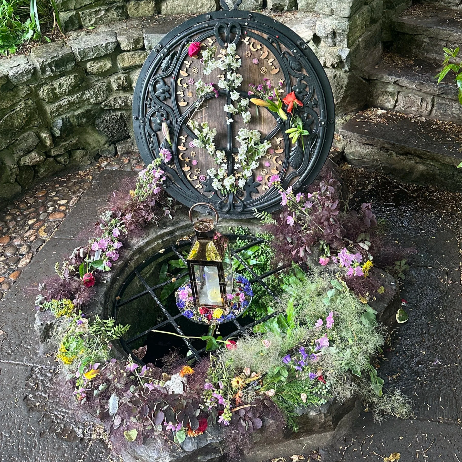 Flower-covered well with a lantern hanging inside, surrounded by flowers and plants, against a stone wall and steps.