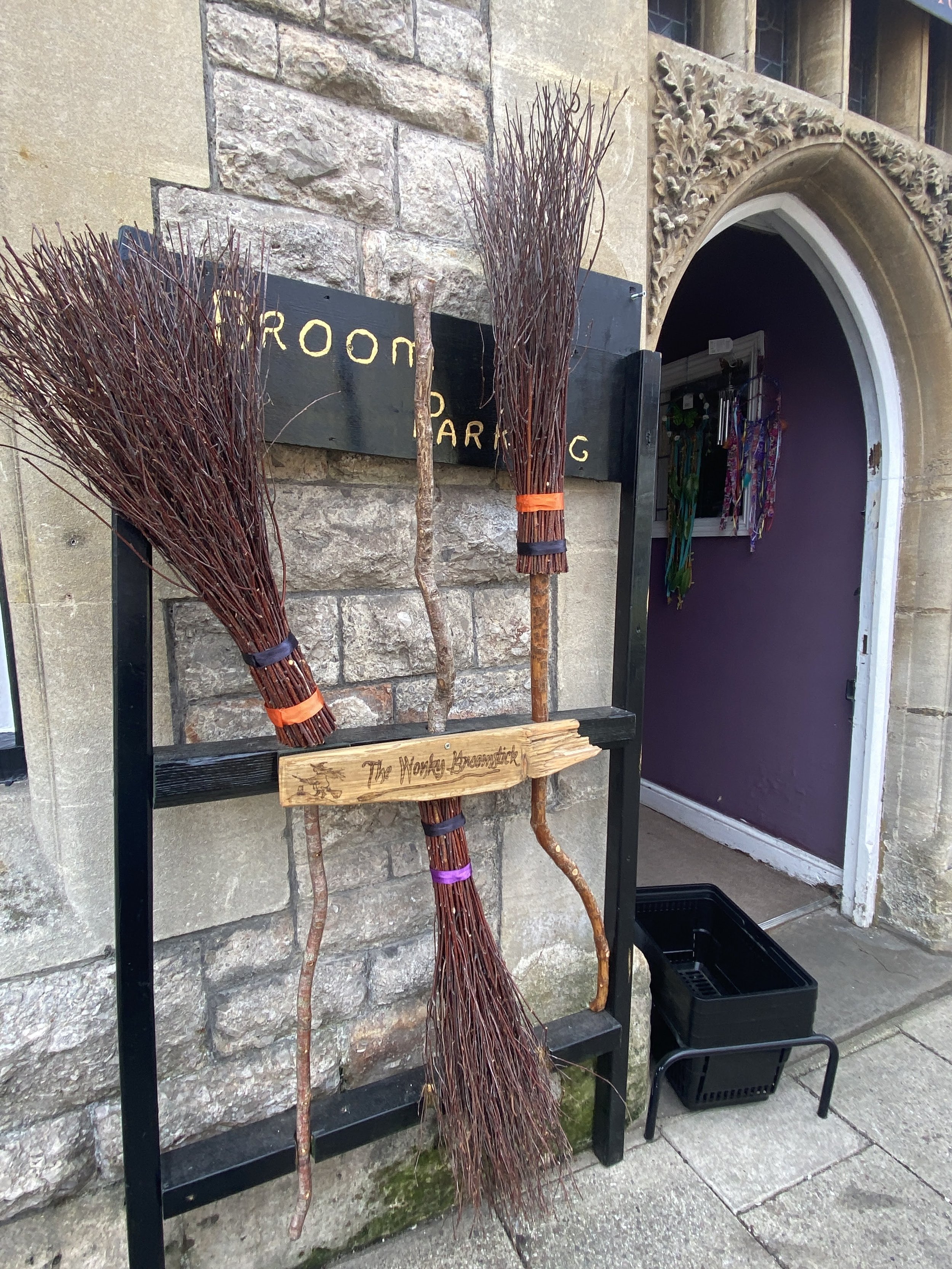 Glastonbury retreat High street. shopping  Three bundles of sticks tied together, leaning against a black display stand outside a stone building with an arched doorway, underneath a sign that reads 'Broom parking'.