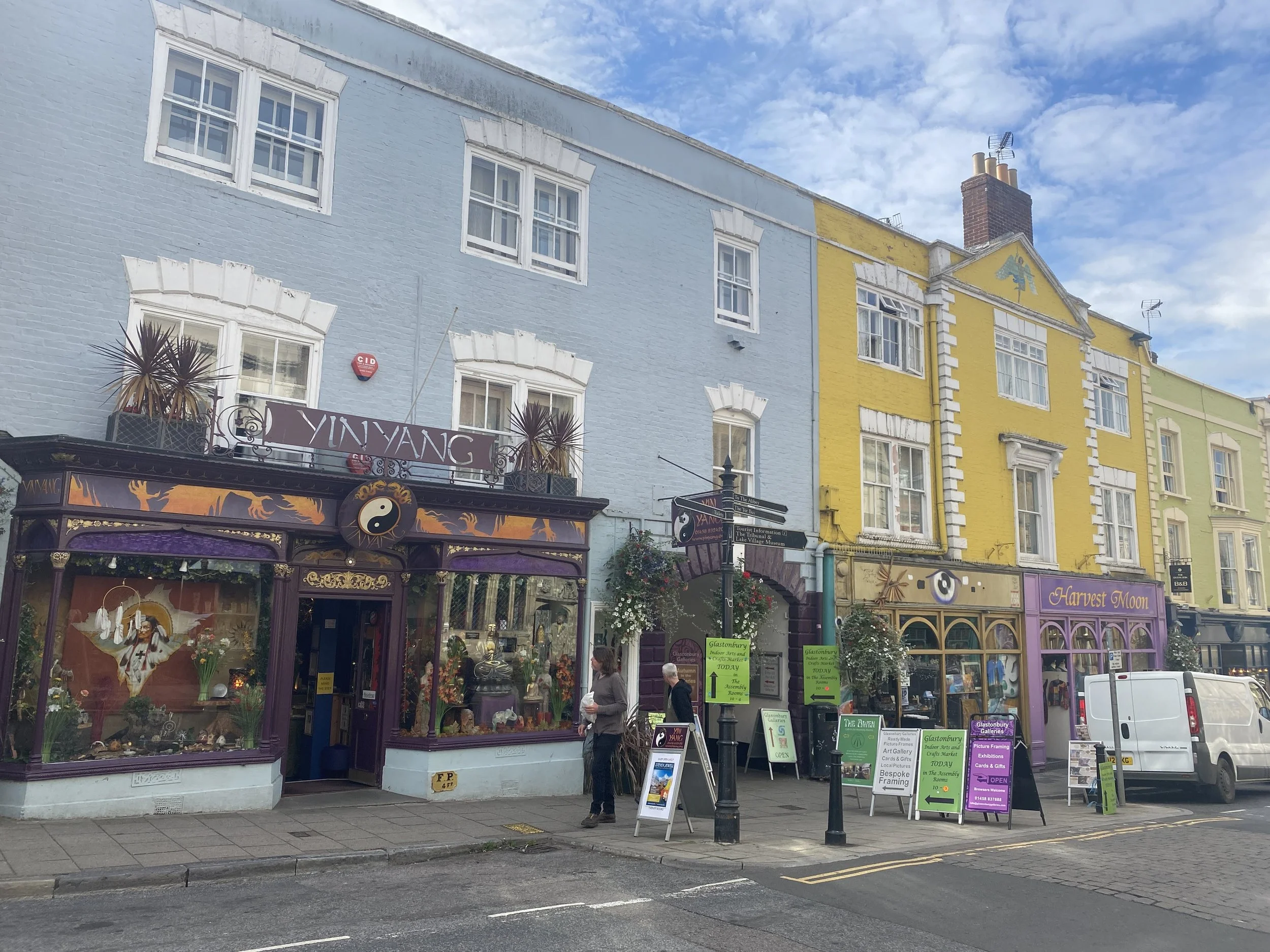 Glastonbury retreat High street. shopping Colorful buildings on a city street, including a blue, yellow, and green building with storefronts and signage, with a cloudy sky overhead.