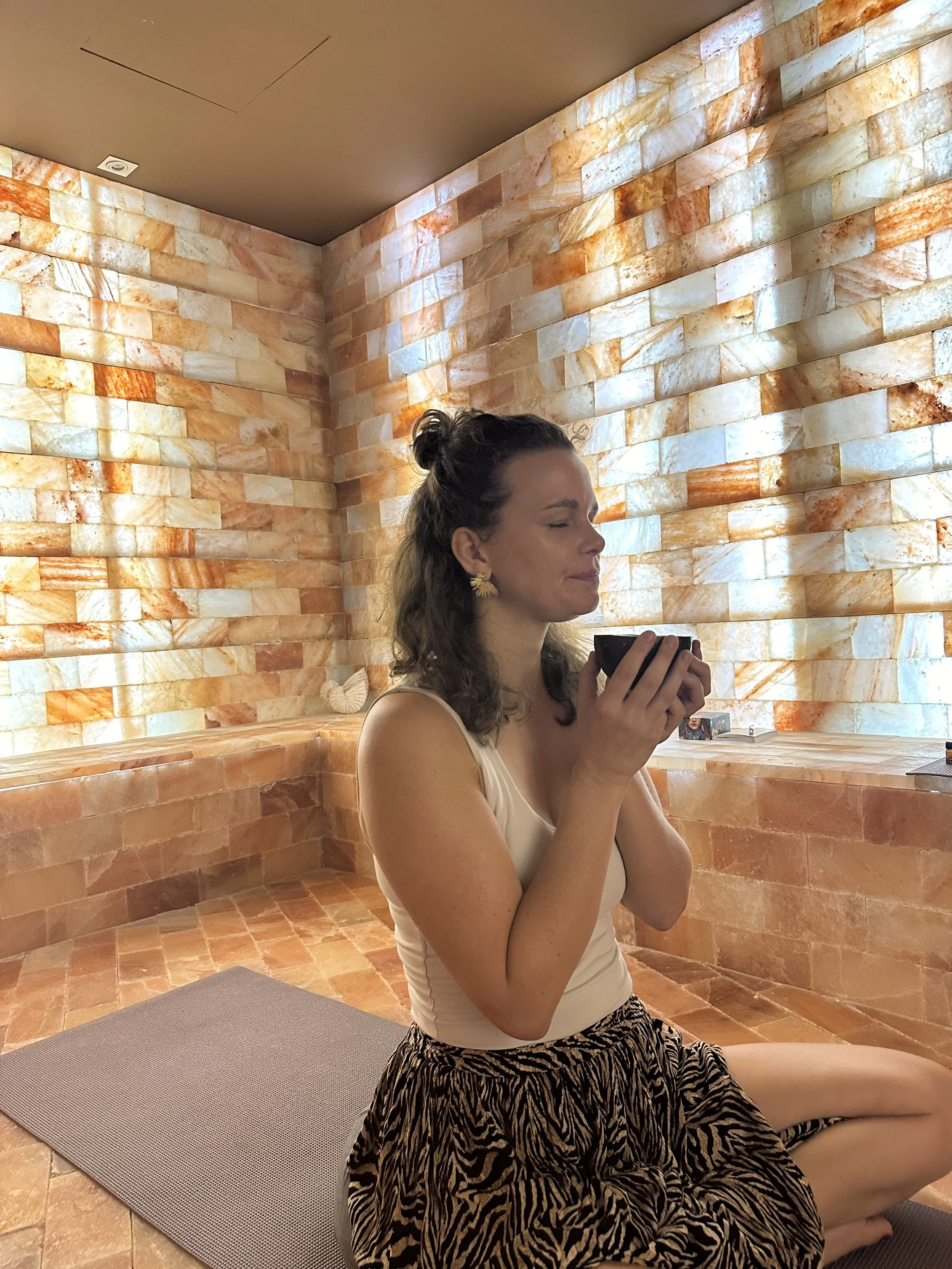 Heike Cushway sitting on a yoga mat in front of a salt wall, holding a cup of Cacao with eyes closed, possibly meditating or relaxing.