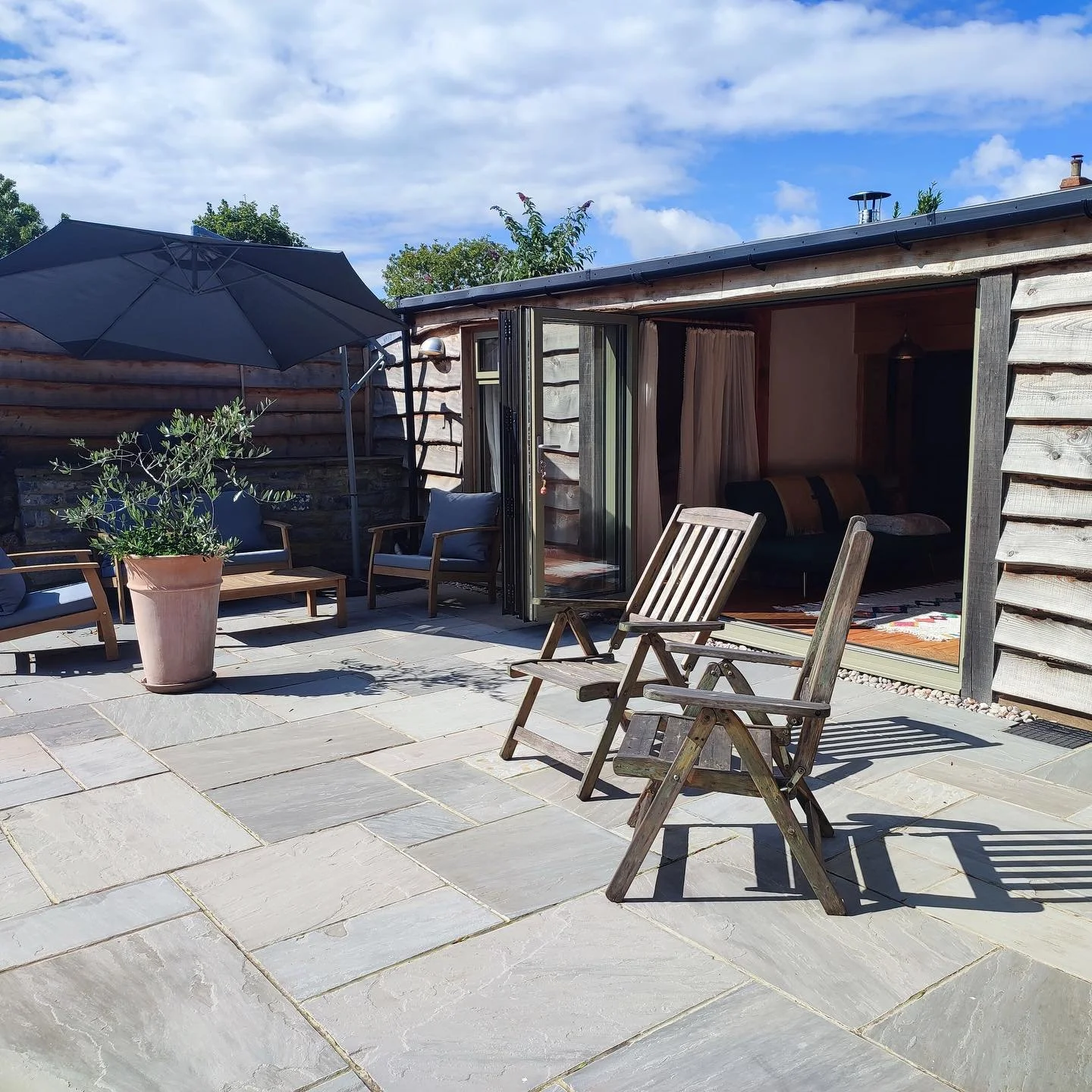 Glastonbury retreat Apothecary Gardens An outdoor patio with wooden chairs, a potted plant, a large black umbrella, and a house with open glass doors, under a partly cloudy sky.