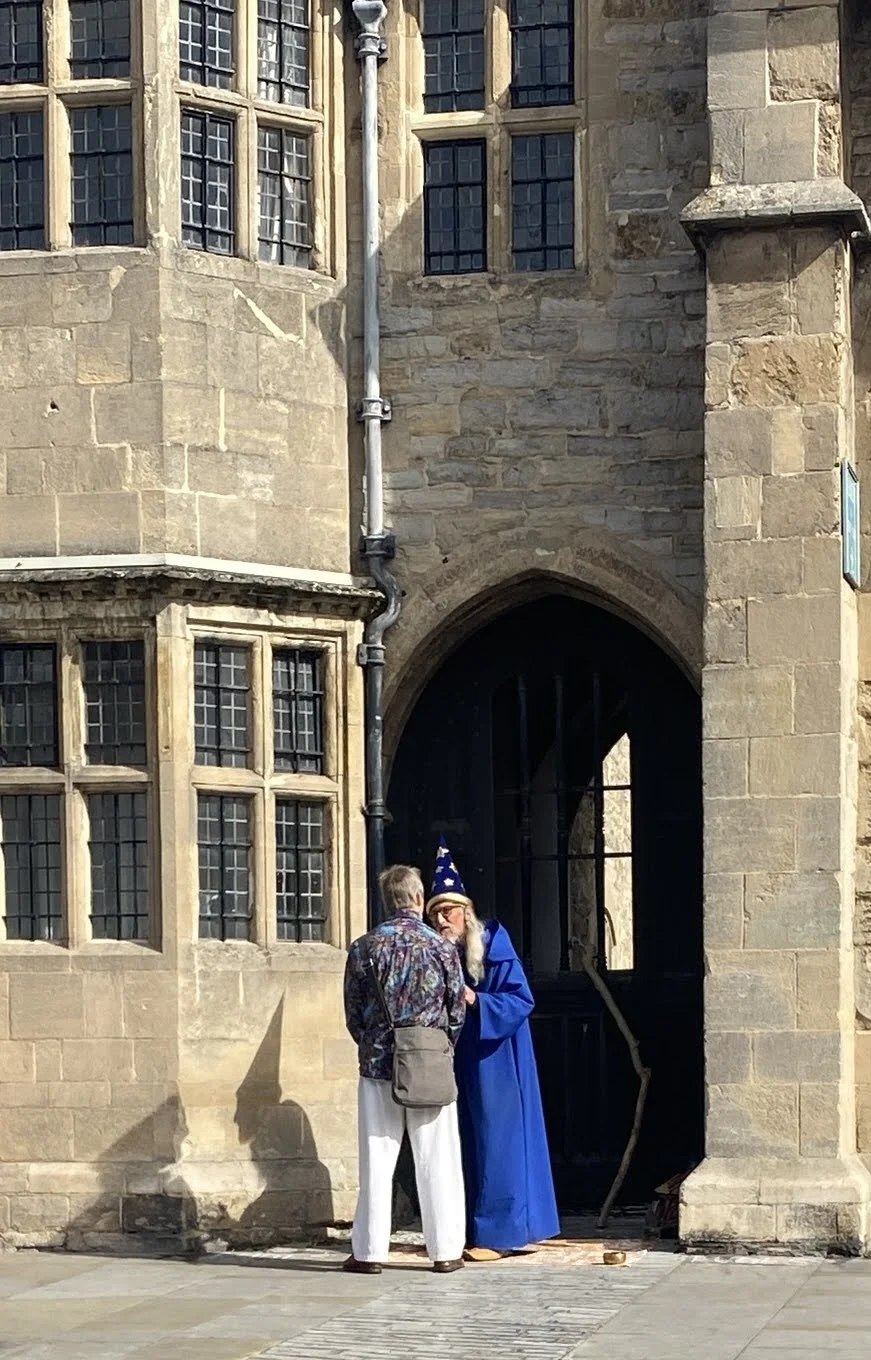 	
Glastonbury retreat High street. s Two people standing outside a historic stone building, one dressed as a wizard with a blue robe and hat, talking to another person in casual clothing.