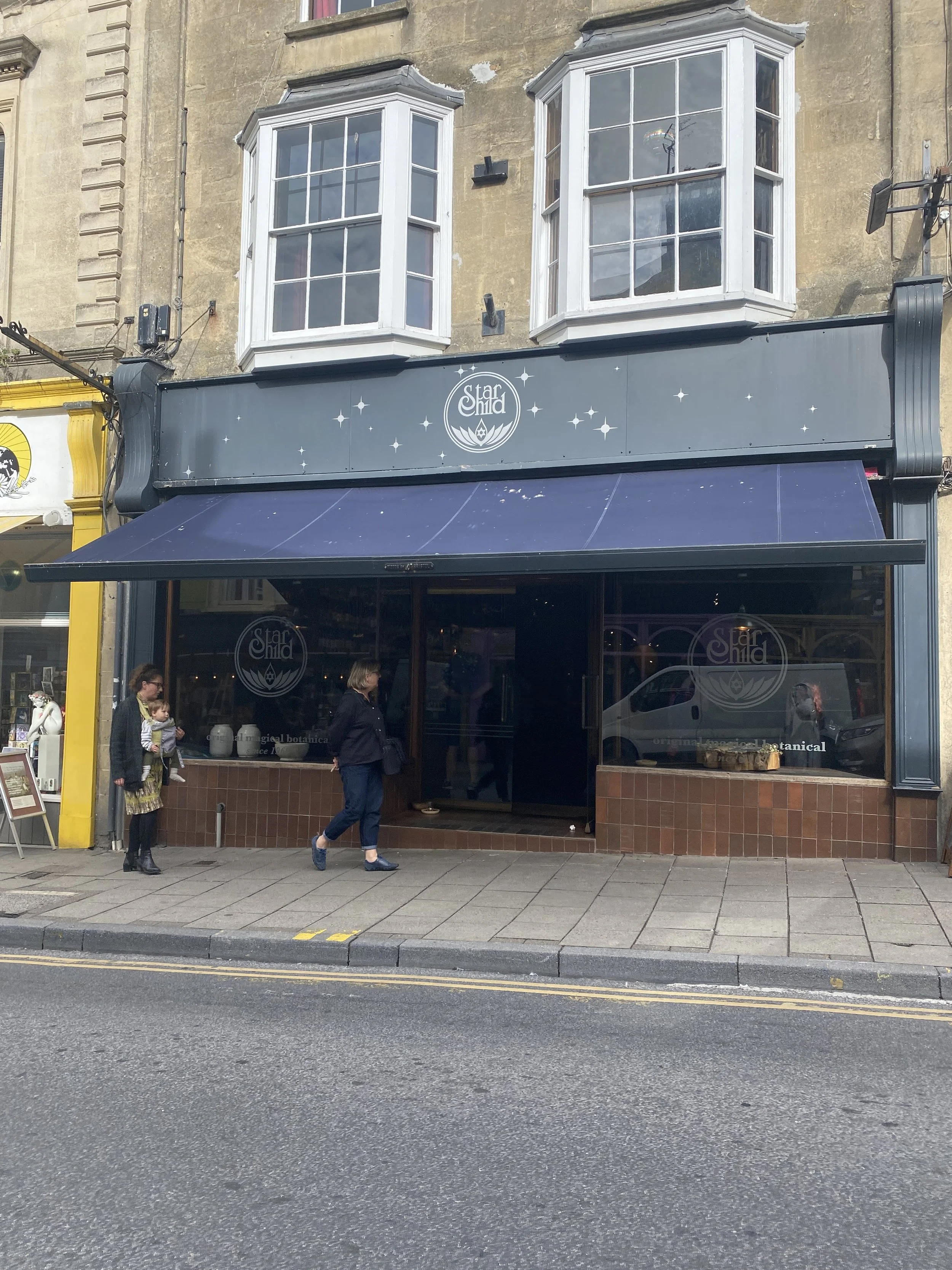 Glastonbury retreat High street. shopping  storefront business named 'Star Child' with a dark-colored awning; large windows with white frames; two bay windows on the second floor; pedestrians walking on the sidewalk in front.