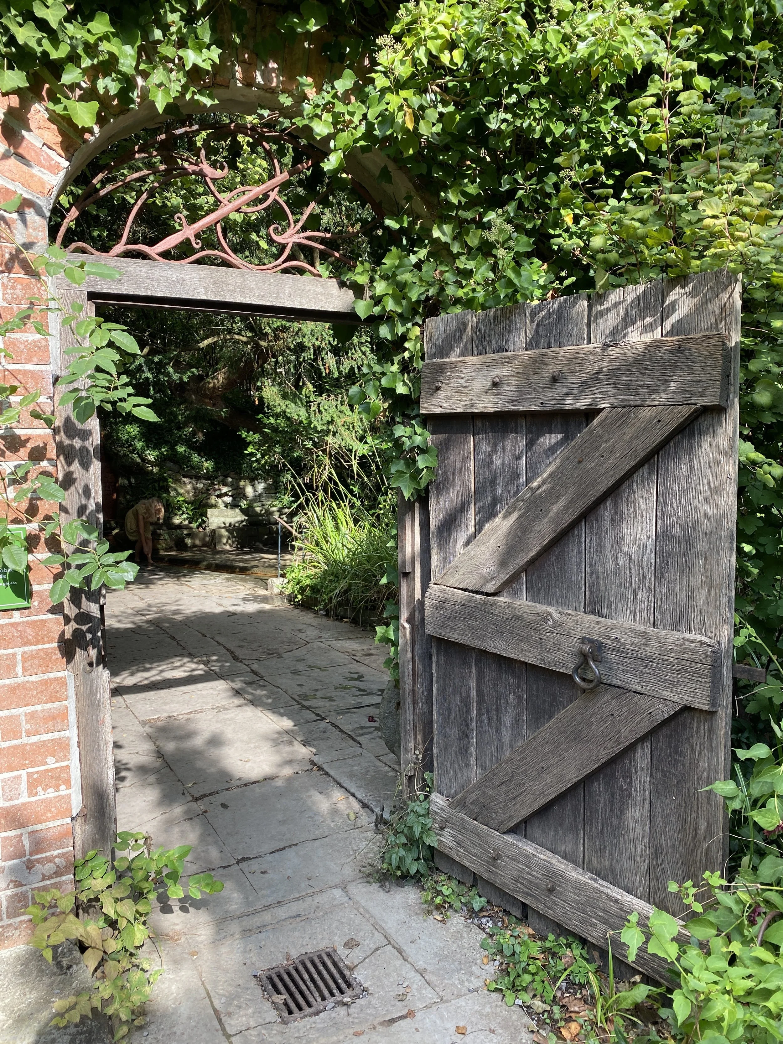 
Glastonbury retreat. Chalice Well A weathered wooden gate partially open, leading into a lush garden pathway with stone paving and greenery, surrounded by ivy and a brick wall.