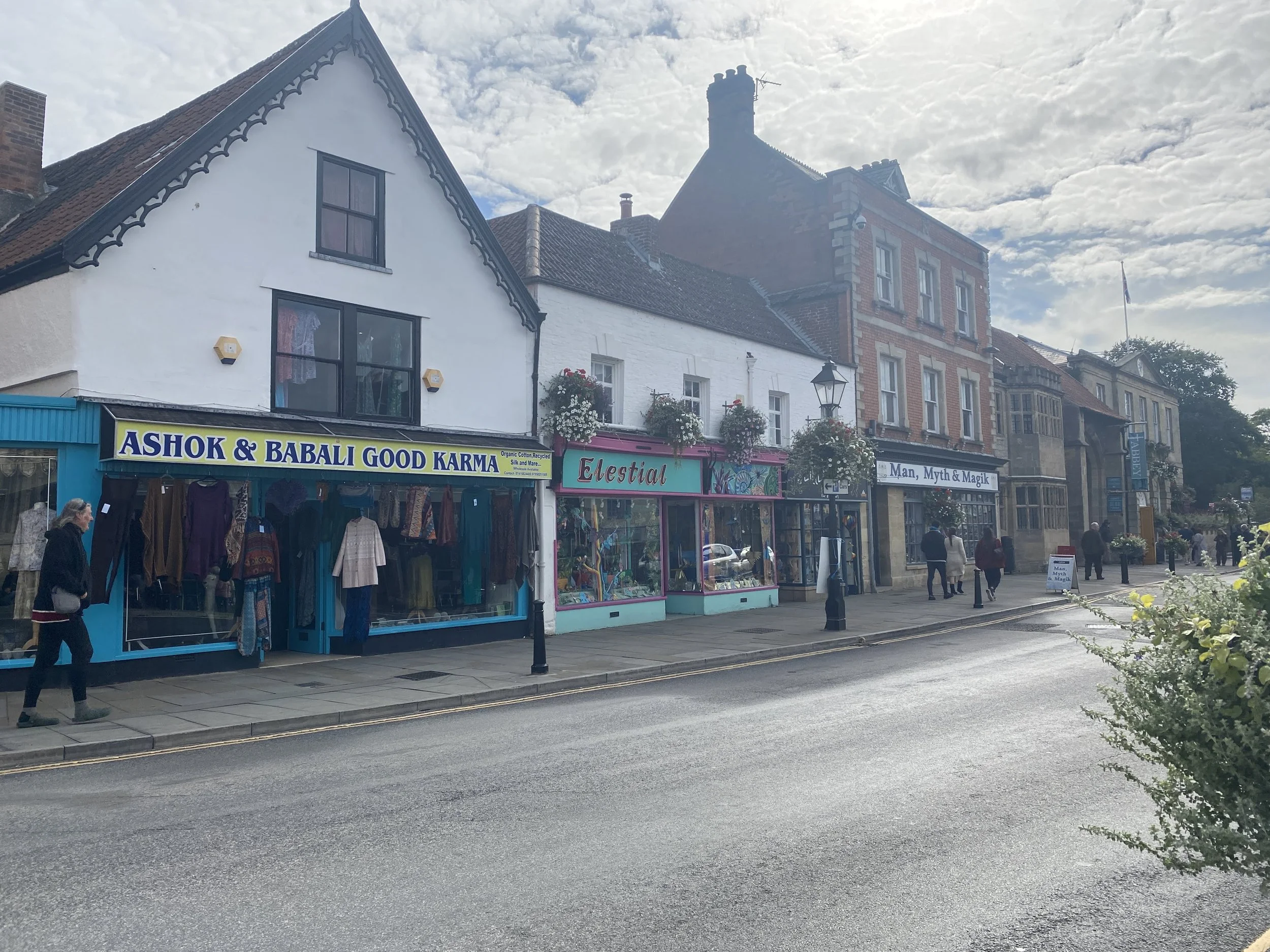 	
Glastonbury retreat High street.  Ashok & Babali Good Karma, with mannequins and clothes in the window, and a store called Elestial with colorful displays. Pedestrians walk along the sidewalk under a partly cloudy sky.