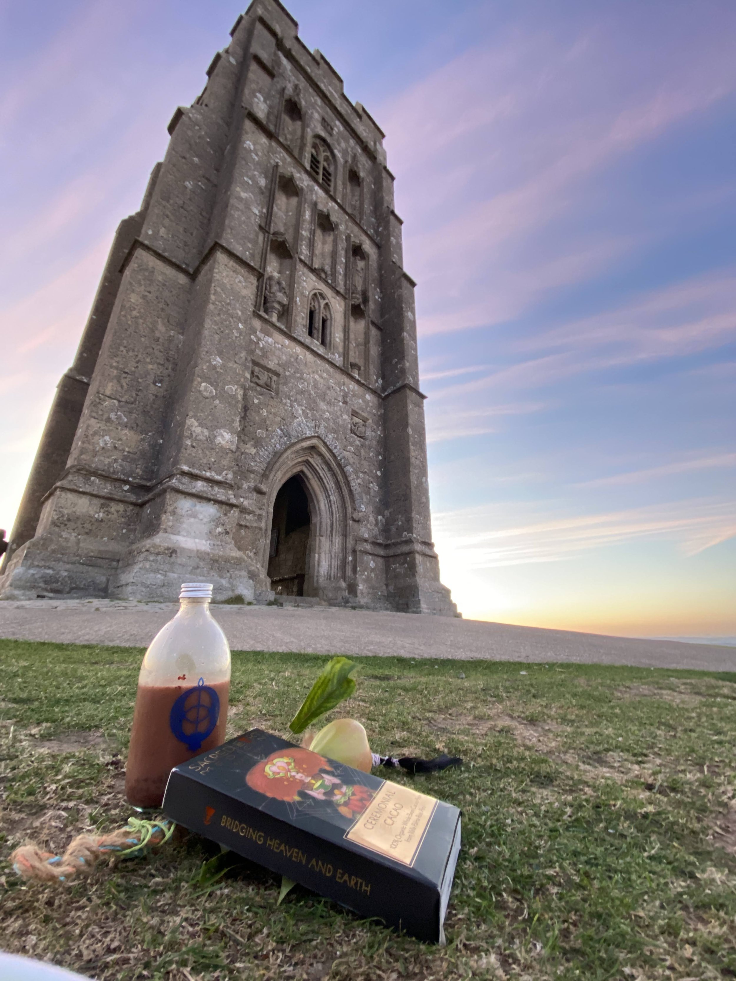 A tall, historic stone tower or church on a grassy hill at sunset, with a bottle of kombucha, a box of ceremonial cacao, an onion, and some small objects in the foreground.