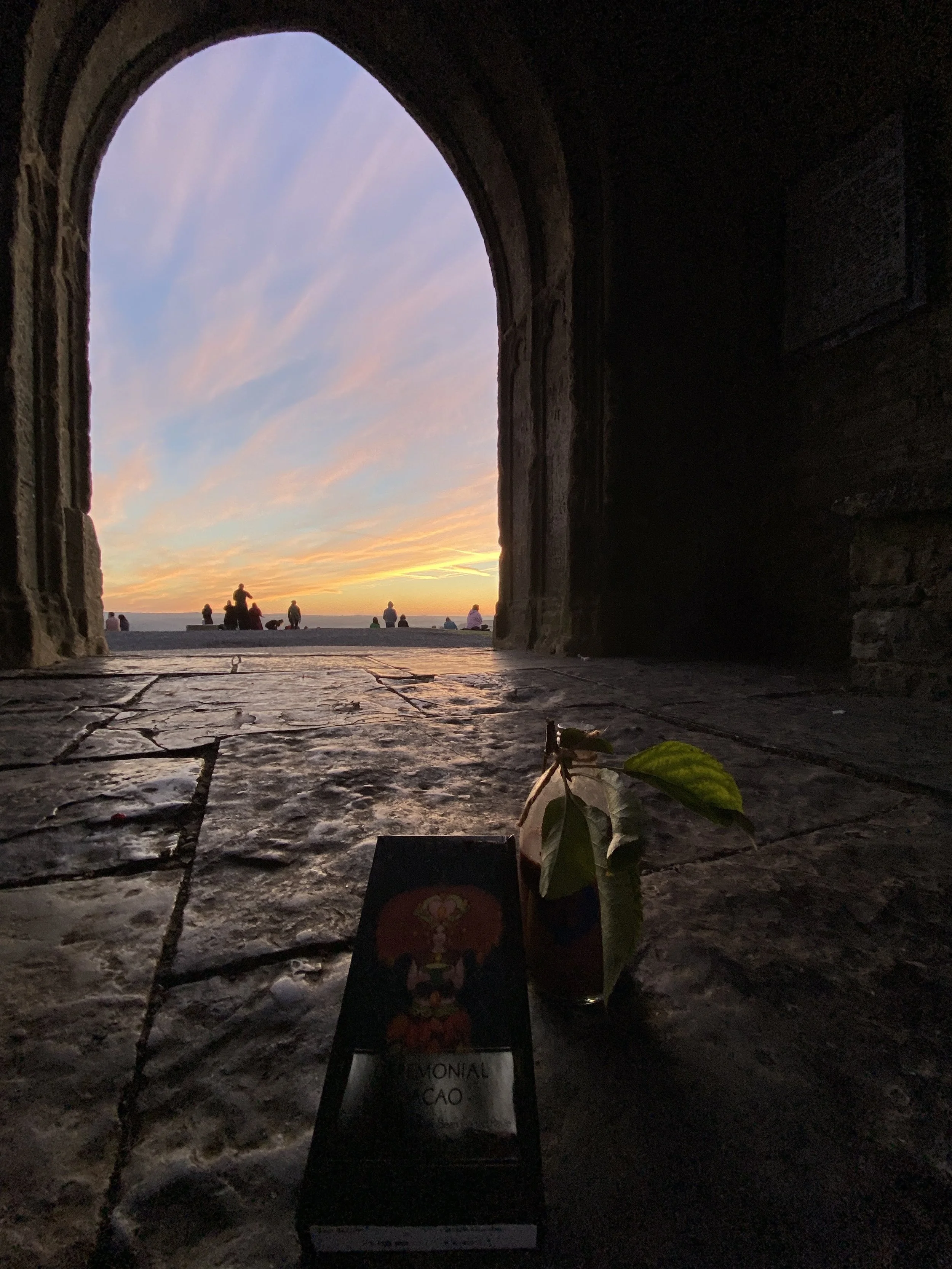 A view from inside Ste Michaels tower, Glastonbury Tor structure looking out through an arched doorway at a sunset over the ocean, with people sitting and standing on the ledge outside. 