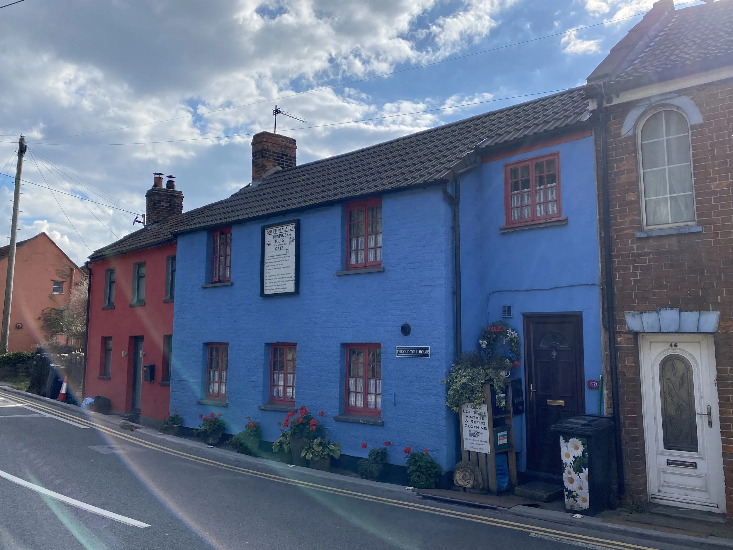Colorful blue and red buildings on a street with flowers outside, under a partly cloudy sky. Glastonbury retreat. High street shopping