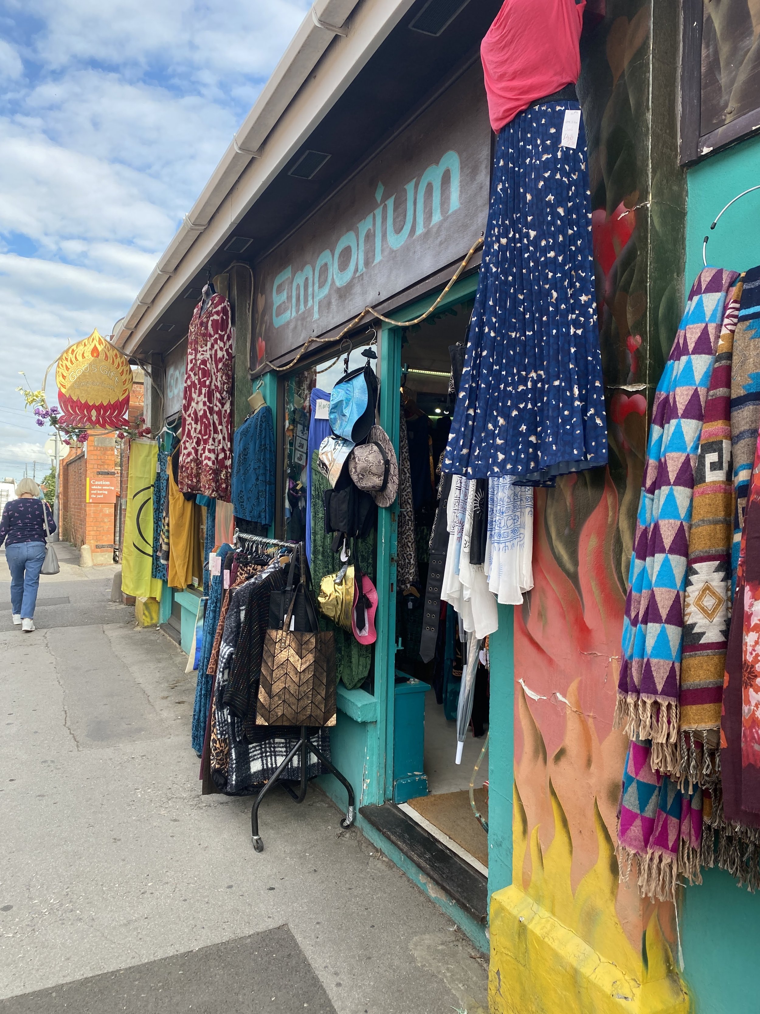 Clothing and accessories display outside a store named 'Emporium' with colorful scarves, dresses, backpacks, and a large decorative lantern, under a partly cloudy sky. Glastonbury retreat High street. shopping