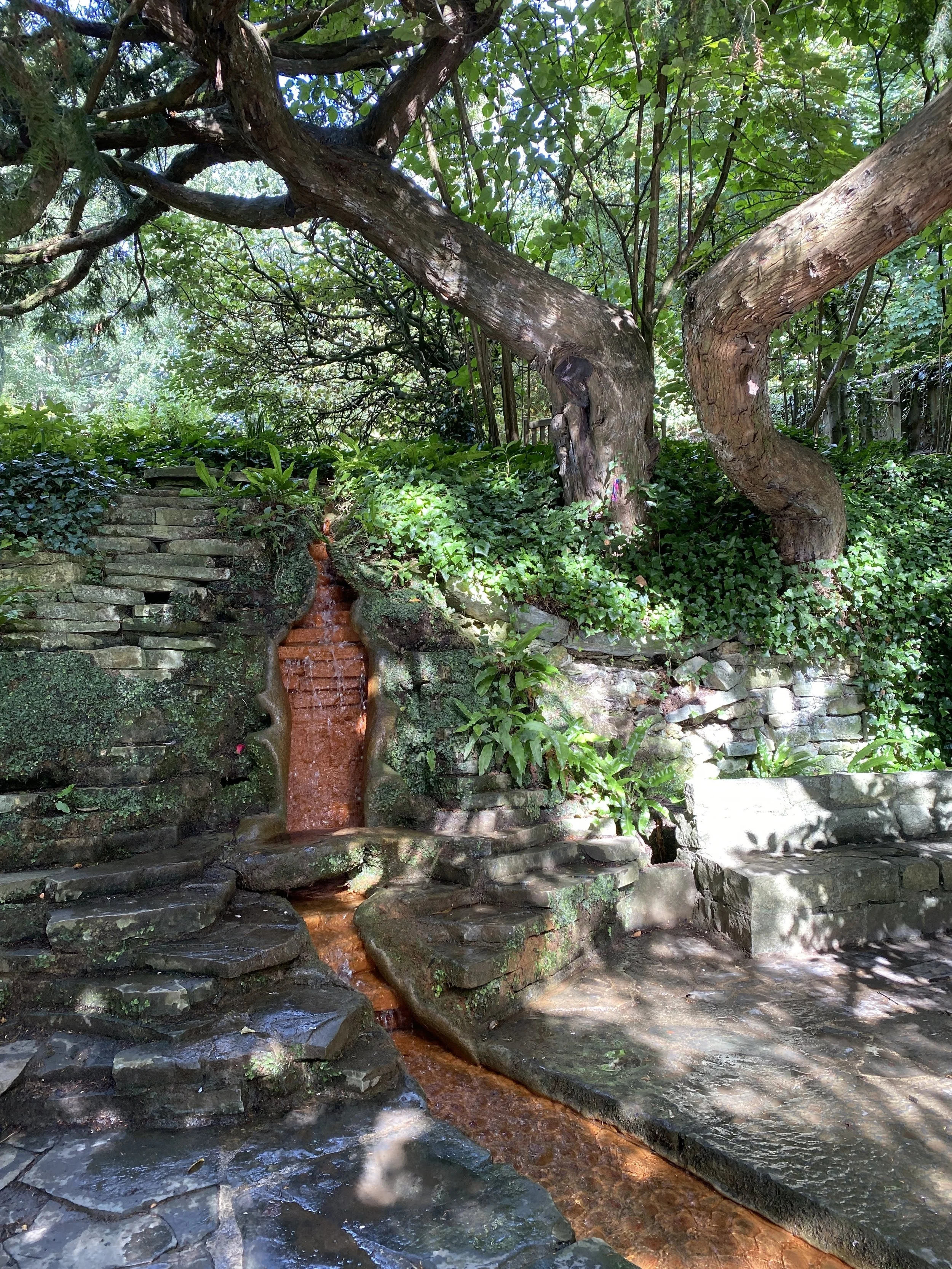 A garden with a small stone waterfall cascading over rocks, surrounded by lush green plants, trees, and a stone seating area in the shade. Glastonbury retreat Chalice Well