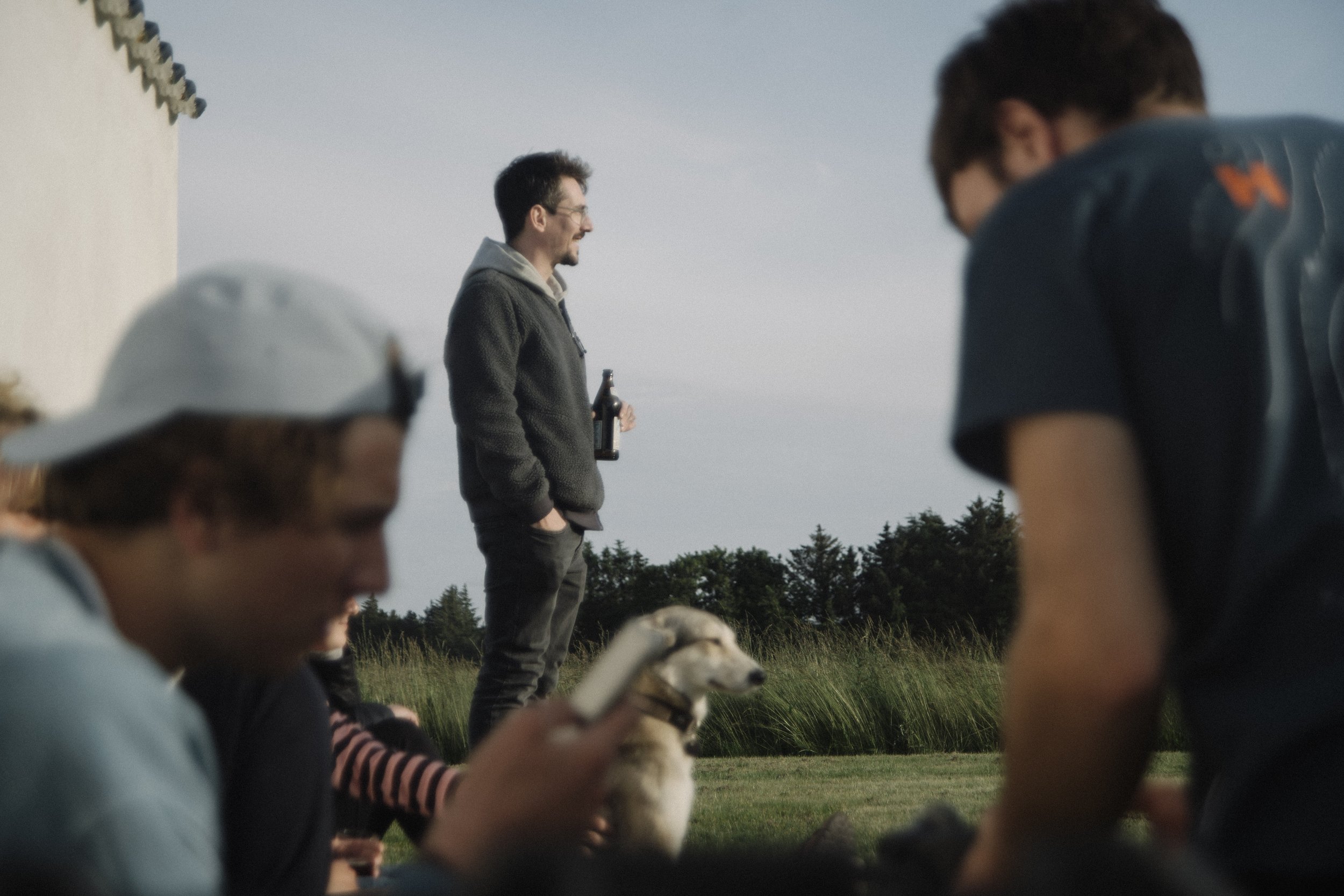 People sitting on the grass outdoors, with one person standing in the background holding a bottle while others look at their phones and a dog sitting on the grass.