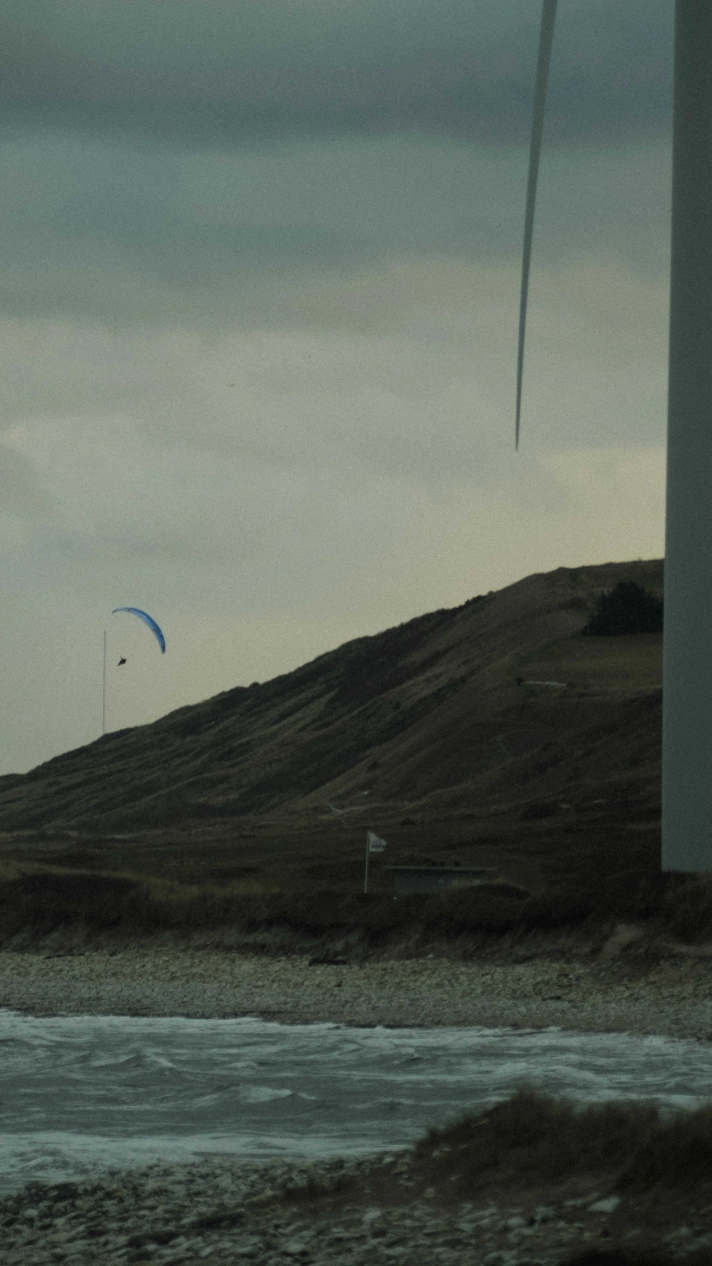 Scenic coastal landscape with grassy hills, a pebble beach, and ocean waves. A paraglider with a blue canopy is flying near a tall tower, and there are two flags on the ground.