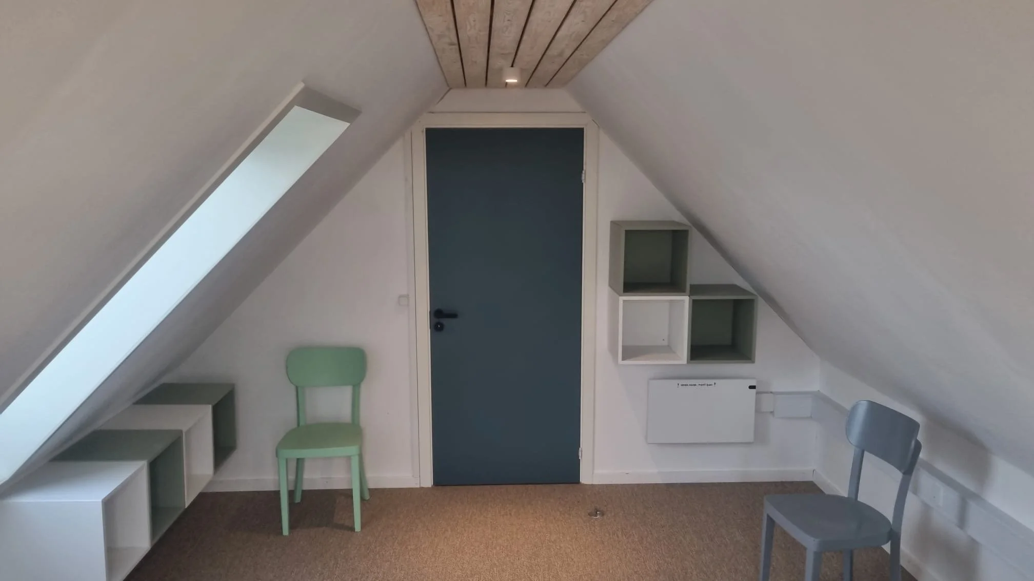 An attic room with white slanted walls and ceiling, a closed blue door centered on the back wall, a green chair on the left, a gray chair on the right, and small white shelves on the right wall. The room has wood paneling on the ceiling and a small w