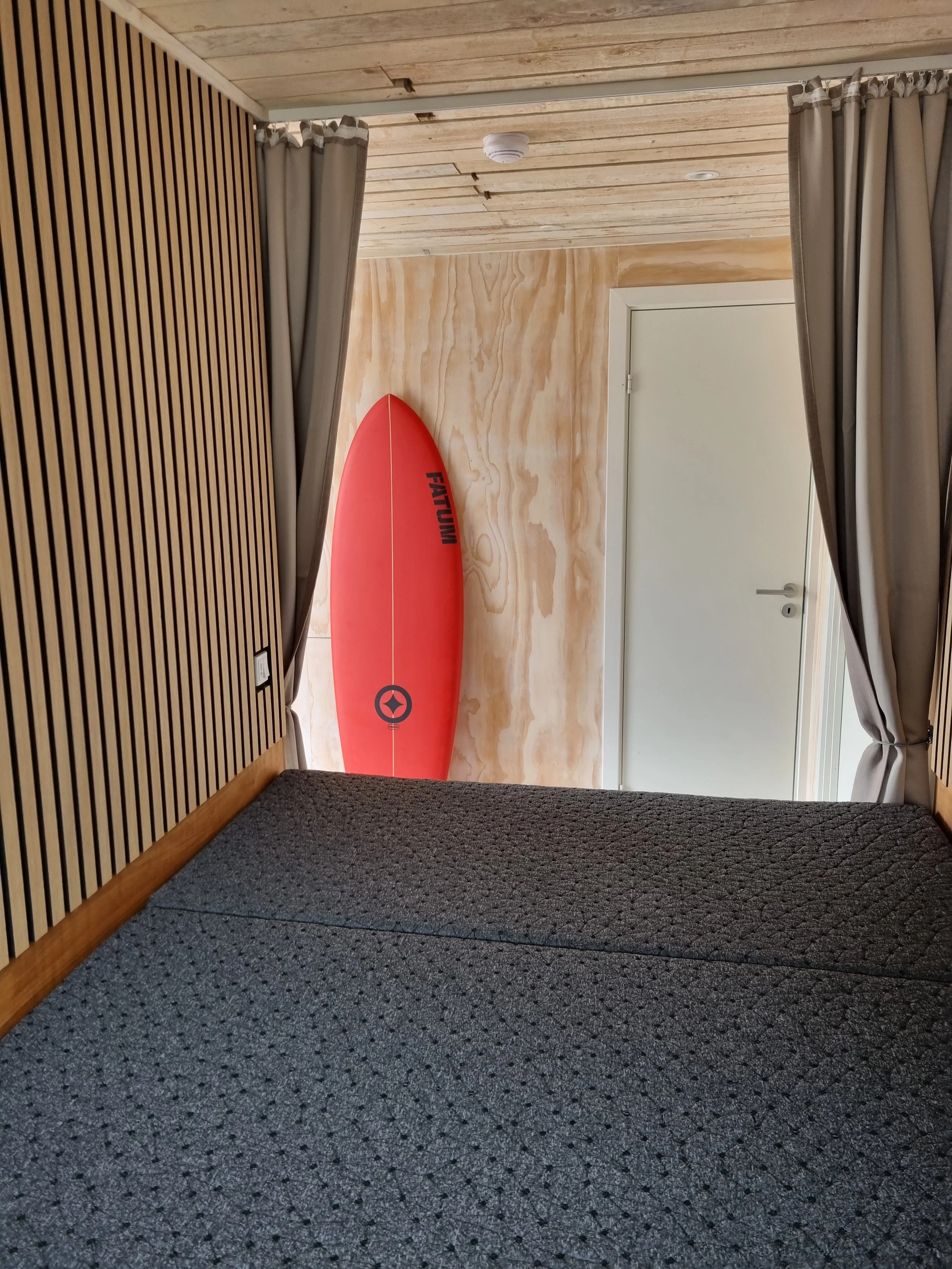 A red surfboard leaning against a wooden wall in a room with beige curtains and a textured gray mattress in the foreground.