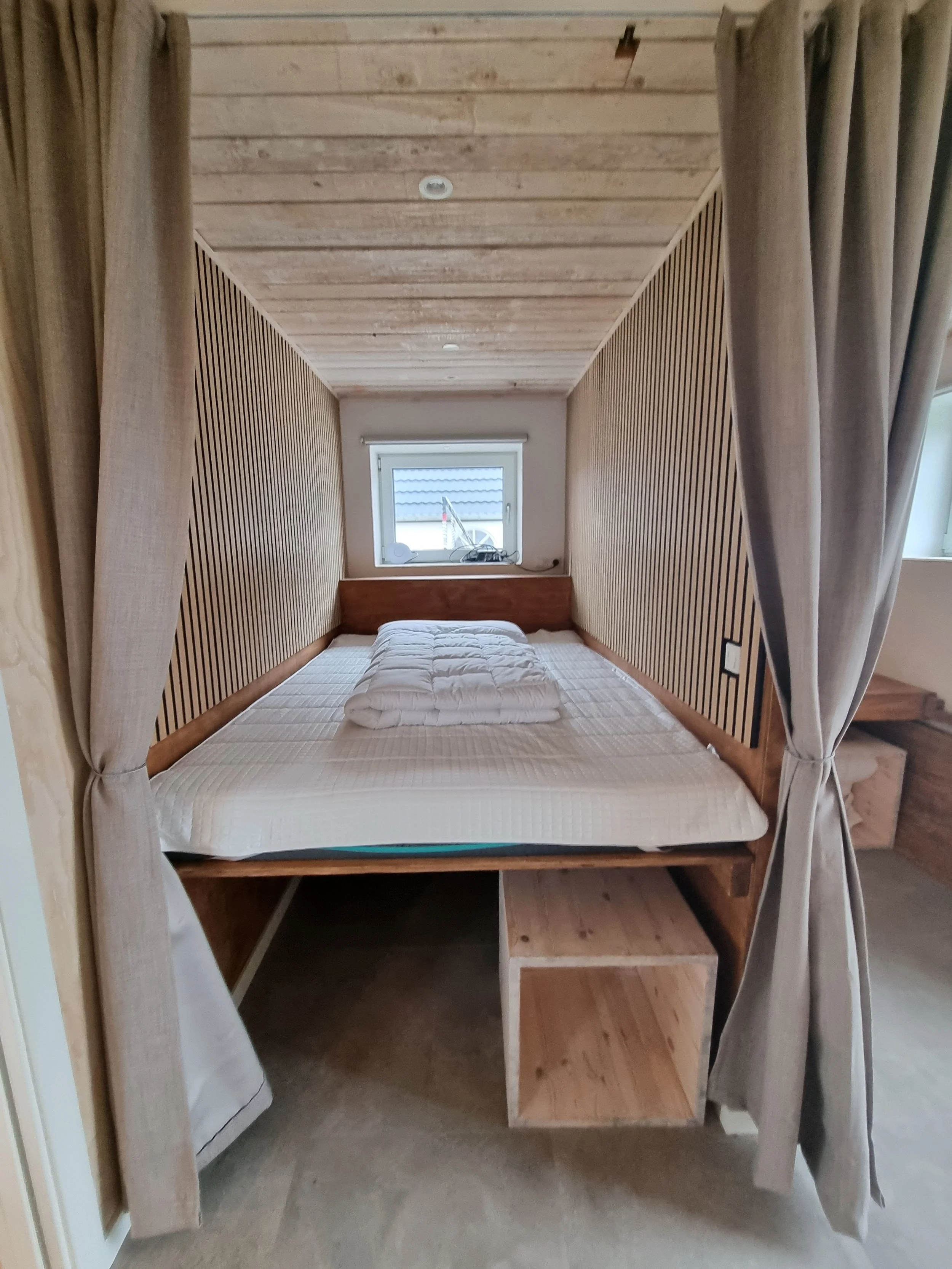 A cozy bedroom with a wooden bed frame and a mattress, partially covered by folded white bedding, under a small window with curtains on either side, in a room with wooden paneling on the ceiling and sides.