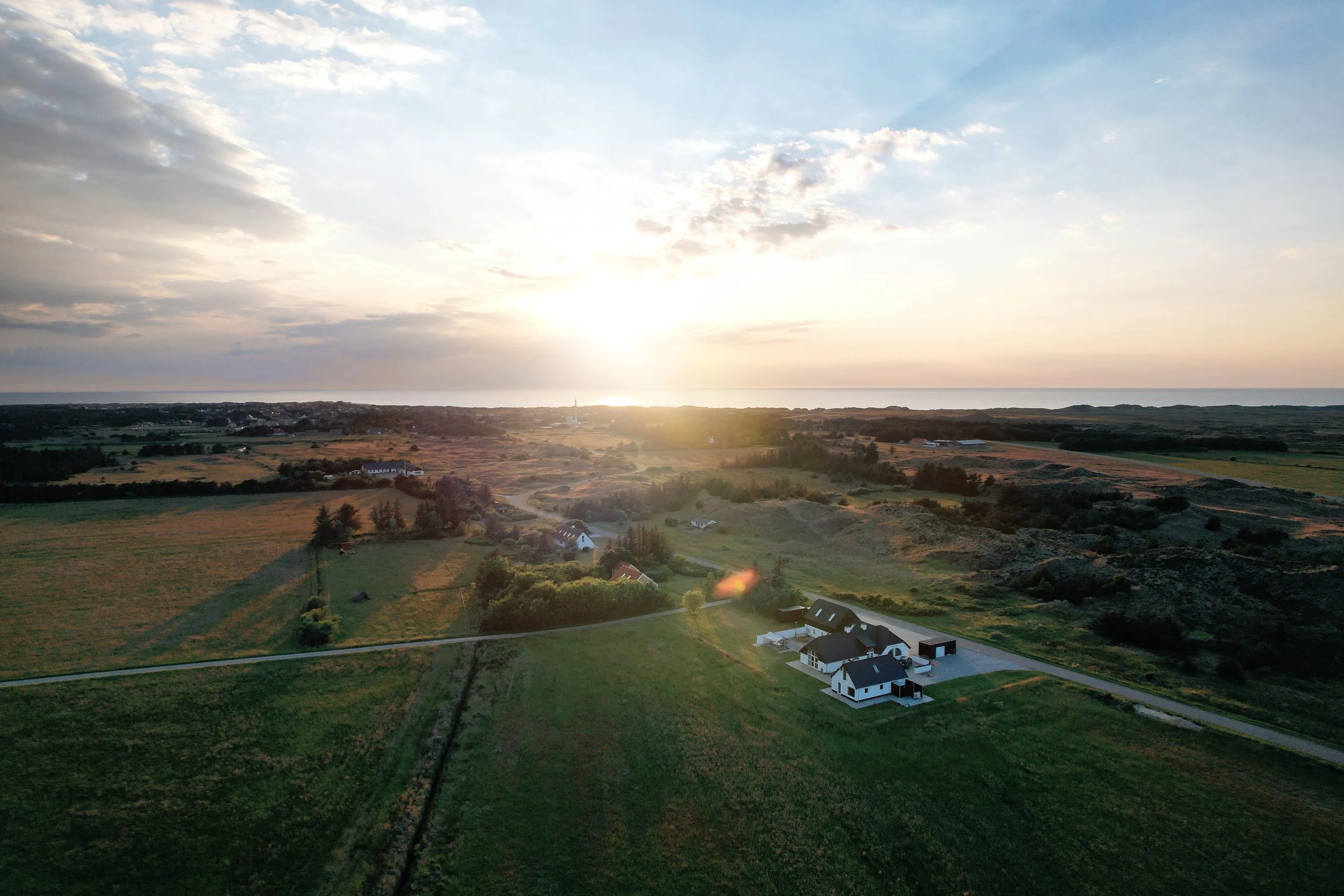 Aerial view of a rural landscape at sunset with fields, trees, a few houses, and a coastline in the distance.