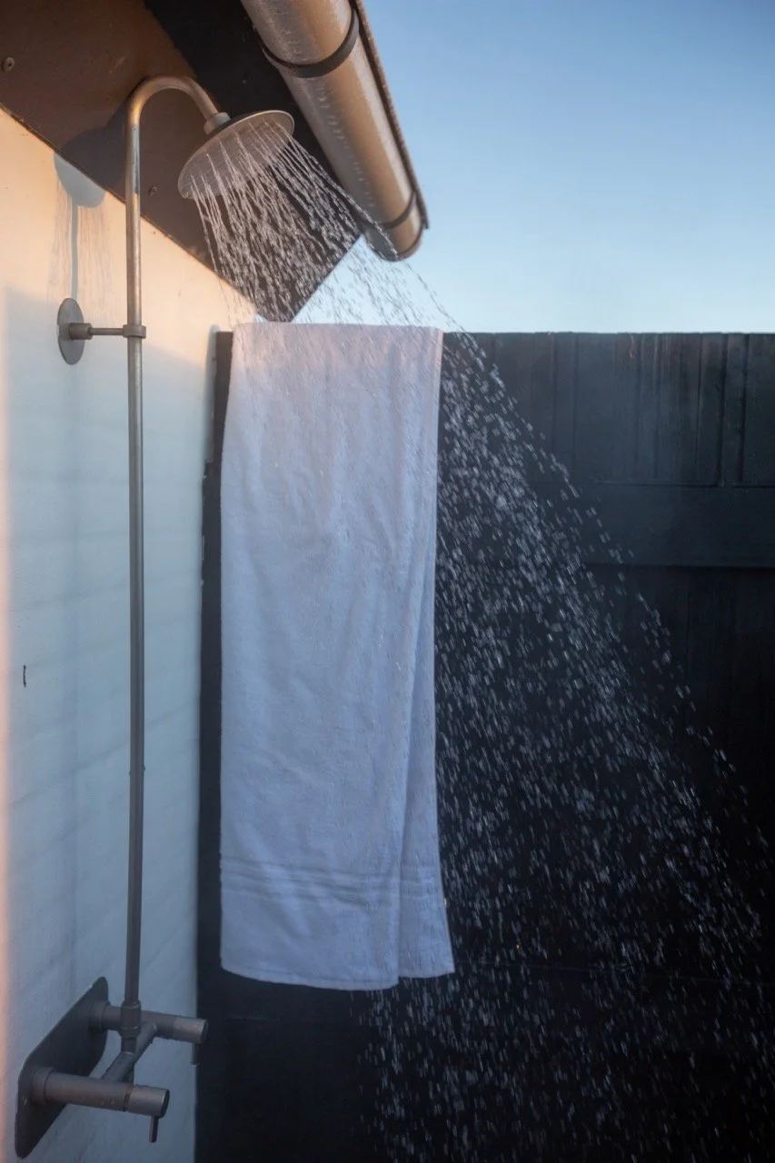 An outdoor shower with water flowing from a round showerhead attached to a pipe, a white towel hanging on a black fence, and a clear blue sky in the background.