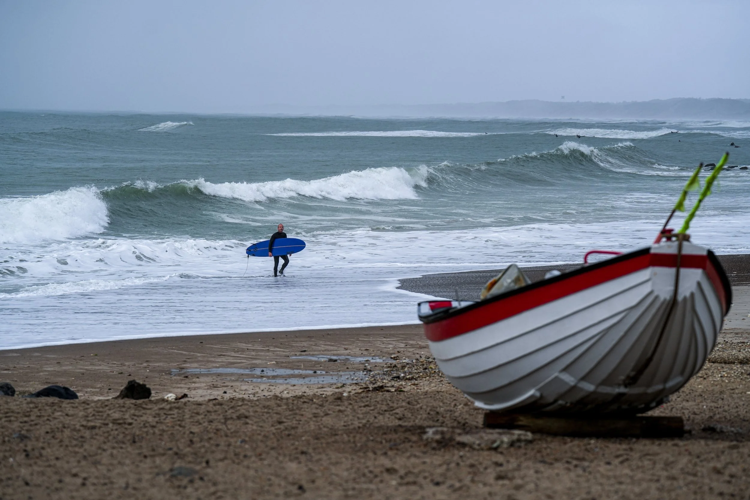 A person holding a surfboard near the shoreline with waves in the background and a boat on the sandy beach in the foreground.
