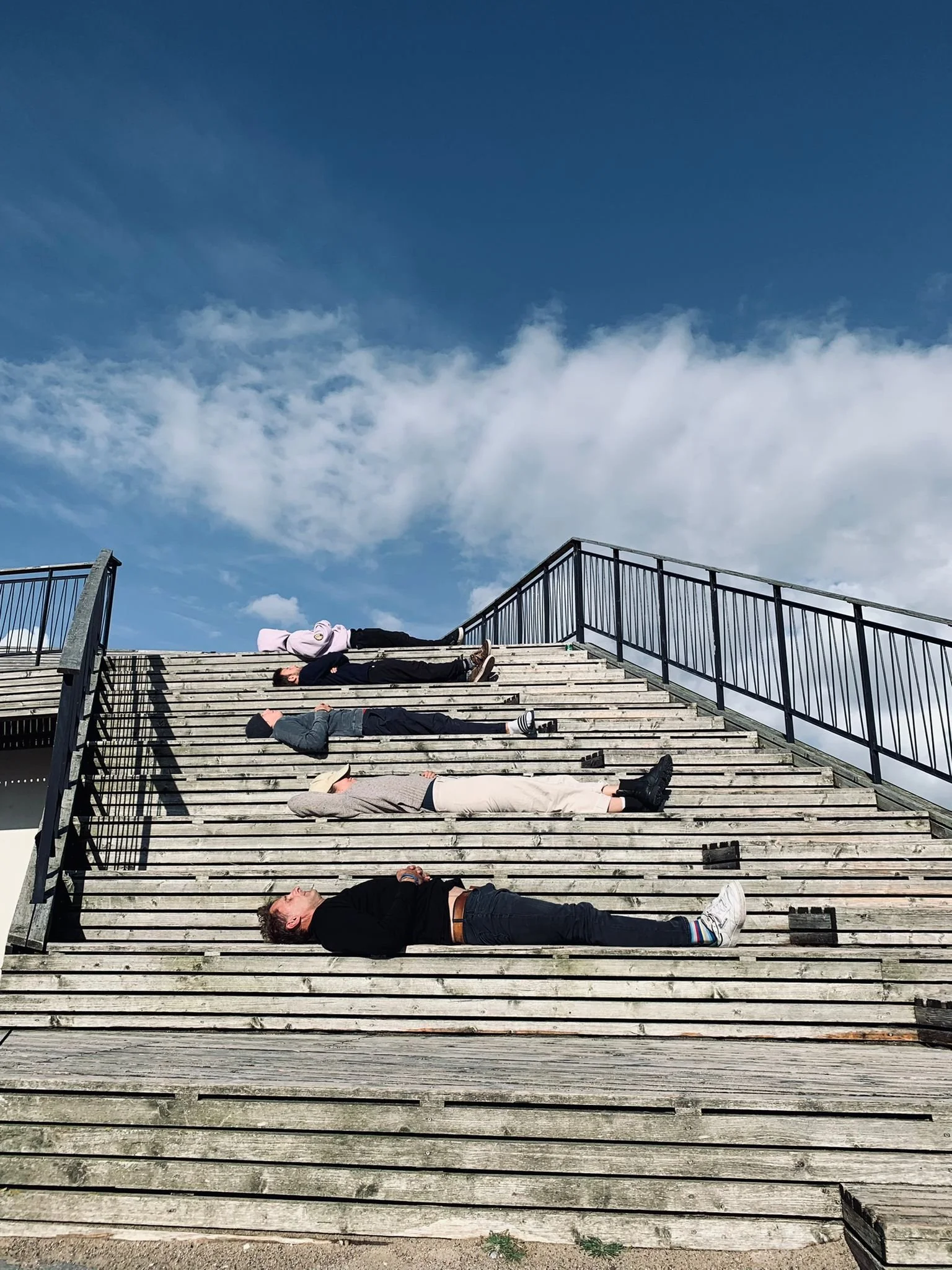 Several people lying on a wooden staircase outdoors under a partly cloudy sky.