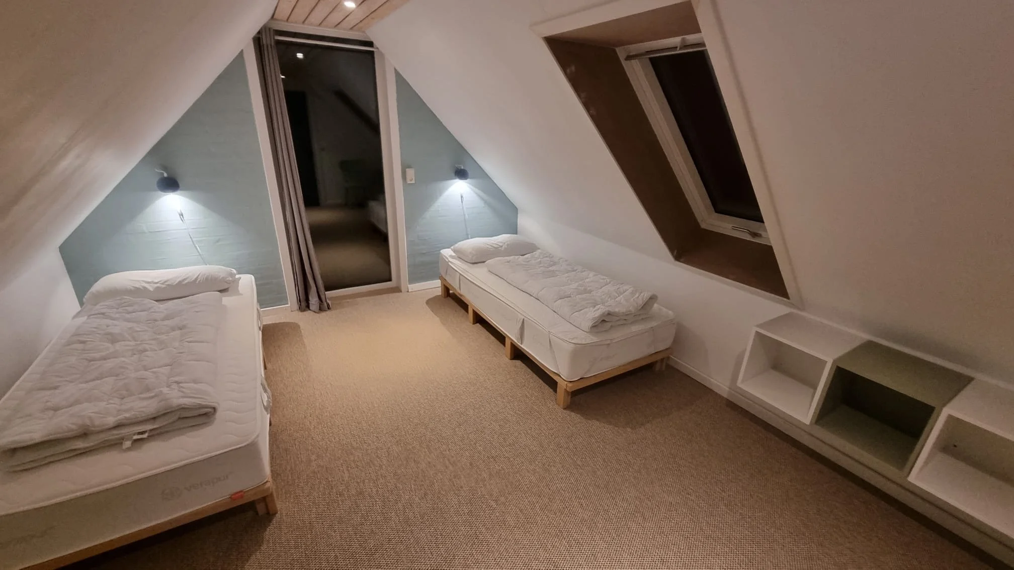 A minimalistic attic bedroom with two single beds, one along the left wall and the other along the right wall. Each bed has a pillow and blanket. There are two wall-mounted reading lights above each bed, and a glass door with curtains leads to a balc