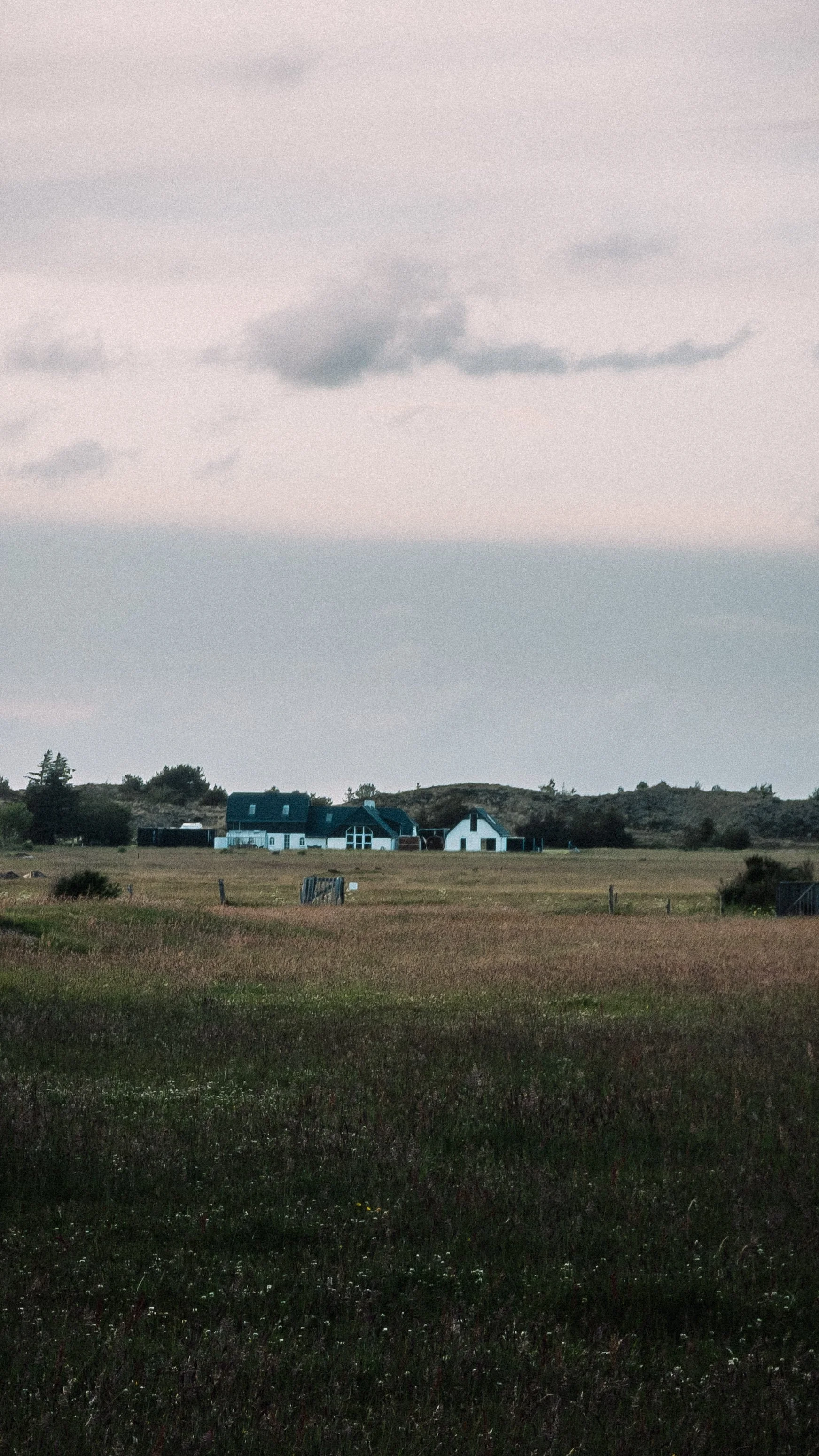 A rural landscape with open grassy fields, some fences, and a large house in the distance under a cloudy sky.
