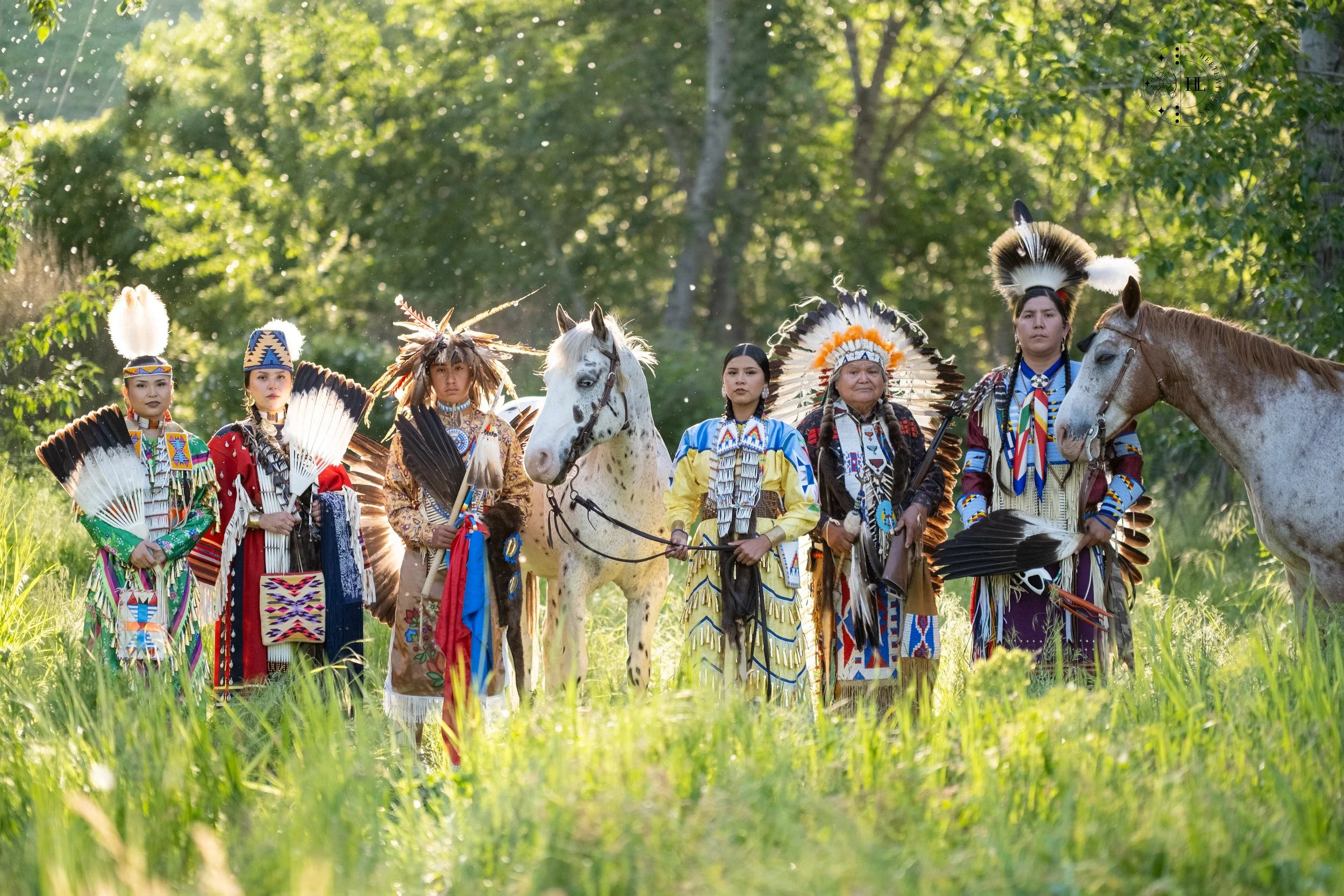 Indigenous dancers in colorful traditional regalia and feather headdresses pose with two horses in a sunlit forest.