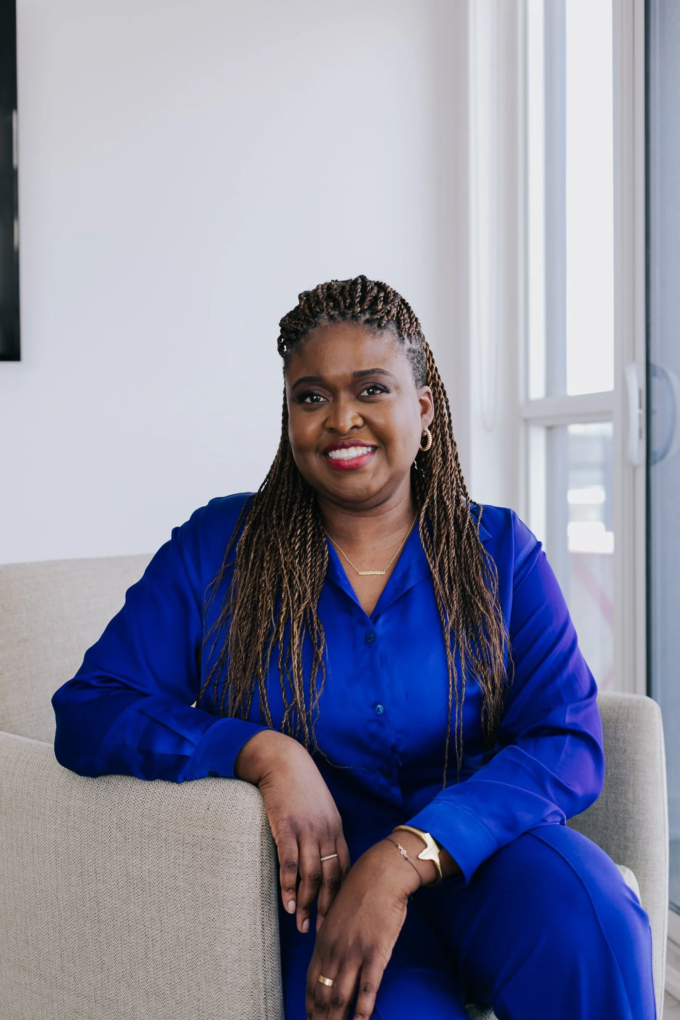 A woman with braided hair, wearing a royal blue blouse, sitting on a beige couch in a bright room with large windows, smiling at the camera.