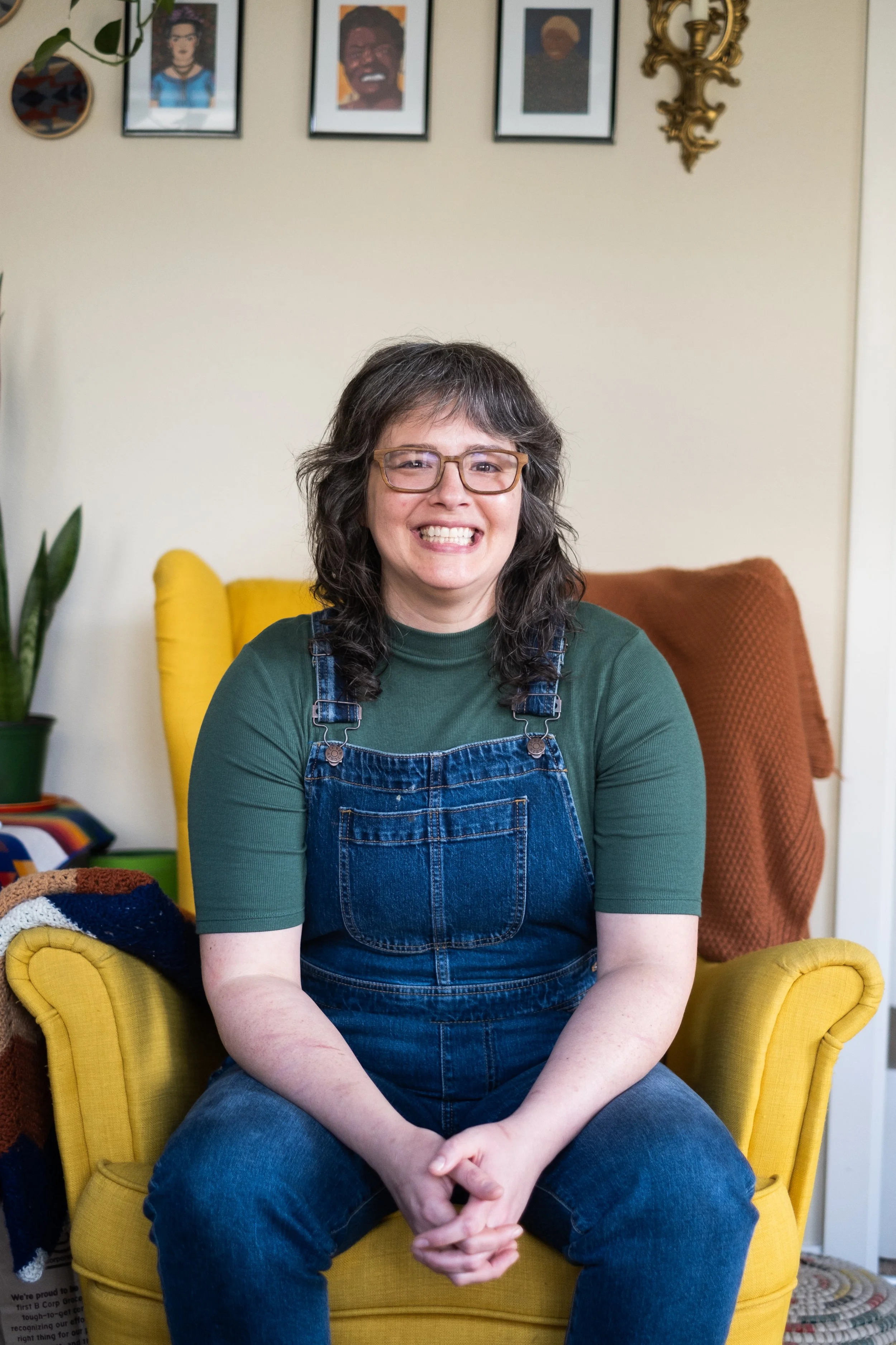 A woman with glasses smiling and sitting on a yellow armchair in a living room with framed artwork and a plant on the wall behind her.