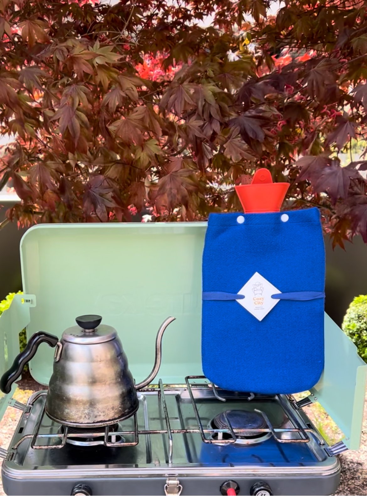 A small camping stove with a silver kettle on it, and a blue insulated bag with a red container sticking out, set against a backdrop of dark red and green leaves.