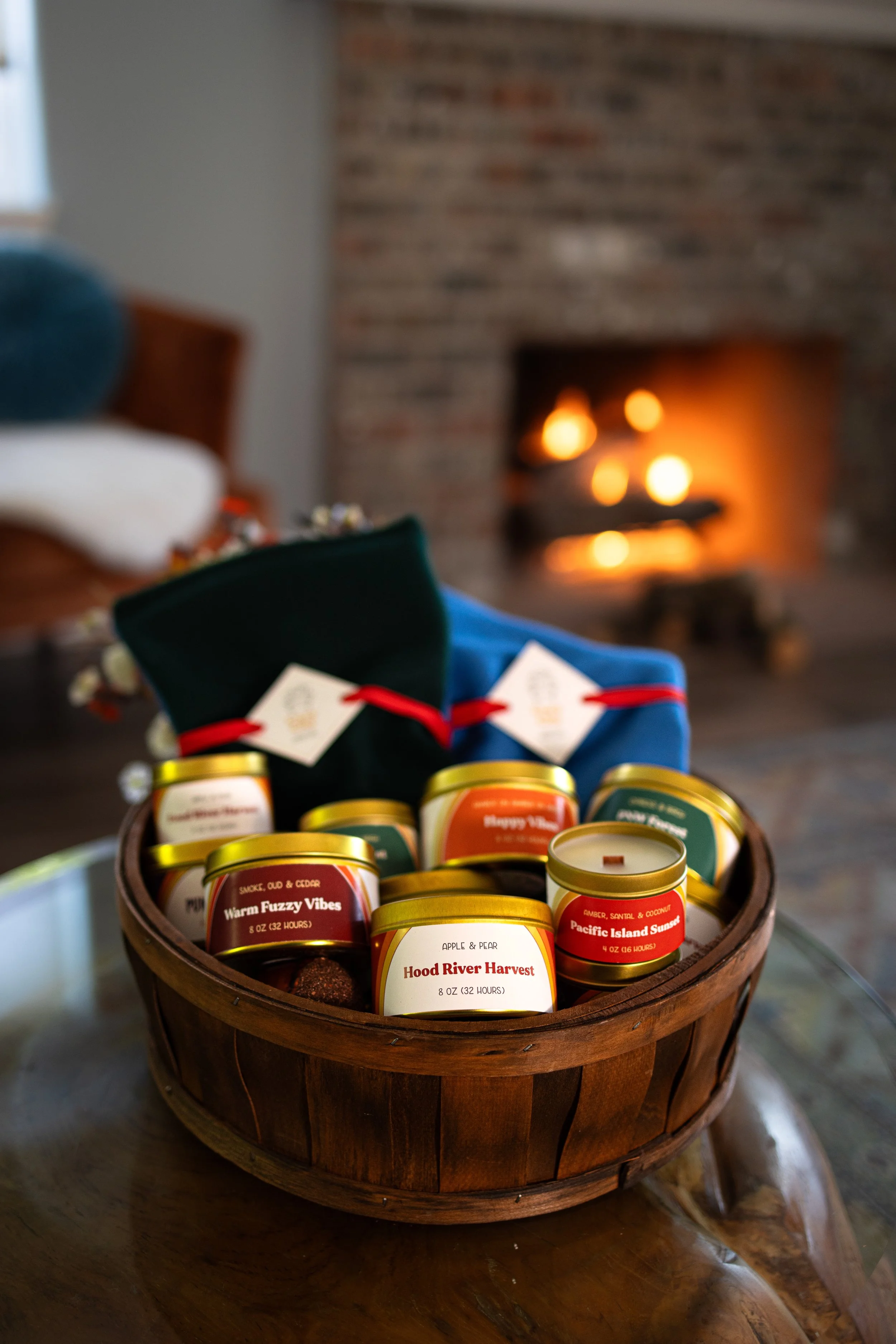 A wooden basket filled with scented candles labeled with various scents, placed on a glass table in front of a fireplace with a warm fire burning.